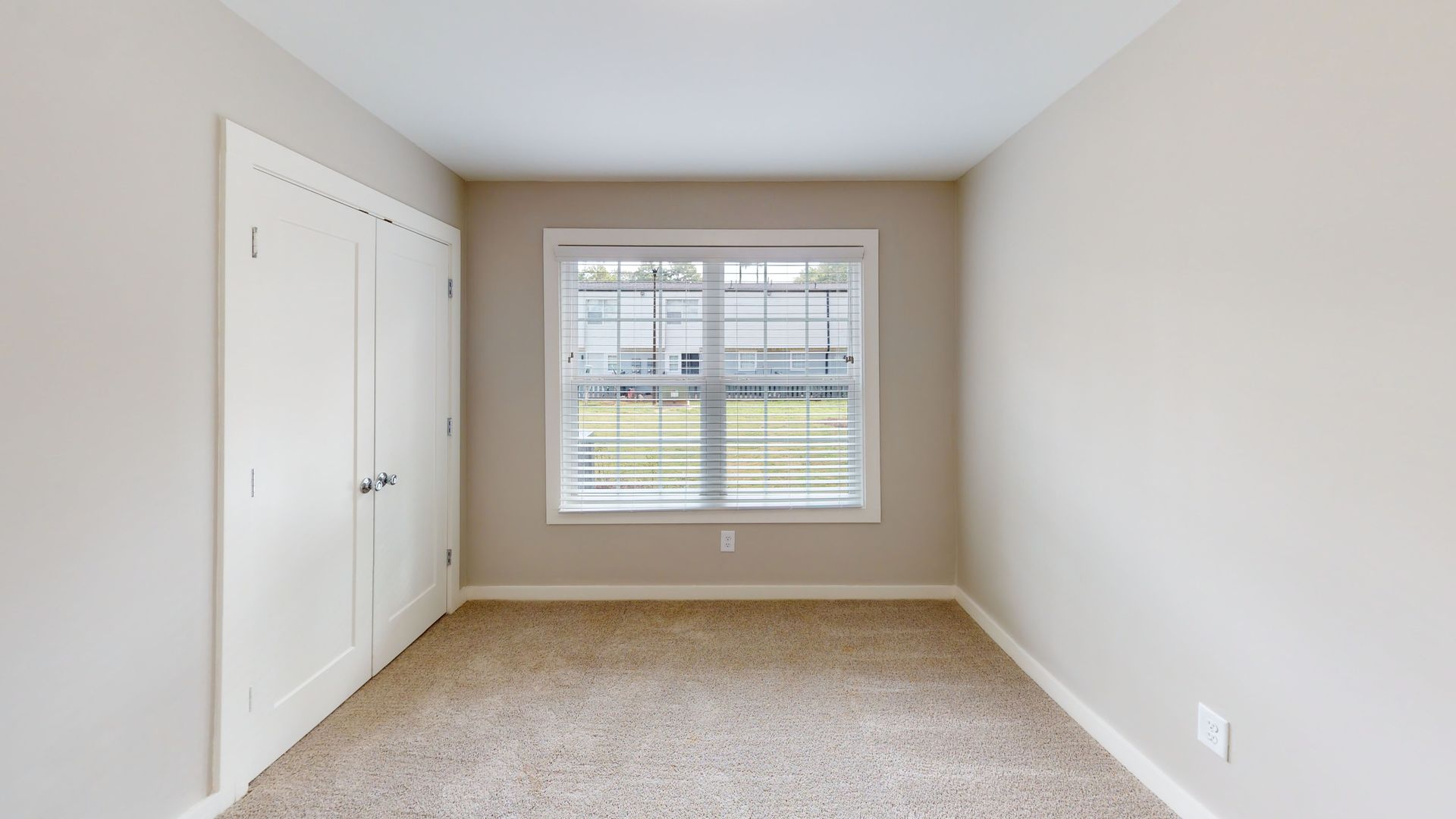 Empty beige bedroom with carpet, closet doors, and a window looking out to a yard.