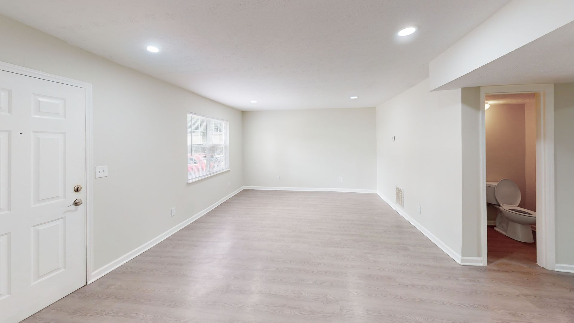 Empty carpeted living room with white walls, recessed lights, a window, and an open doorway to a bathroom.