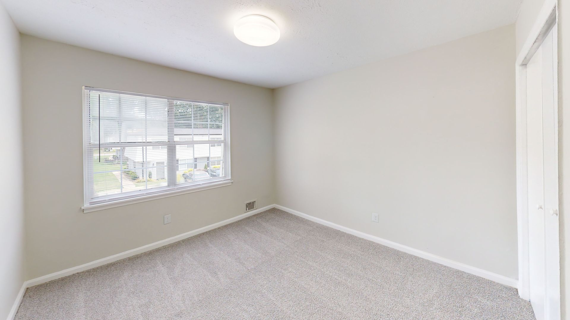 Empty beige bedroom with carpet, window blinds, and ceiling light