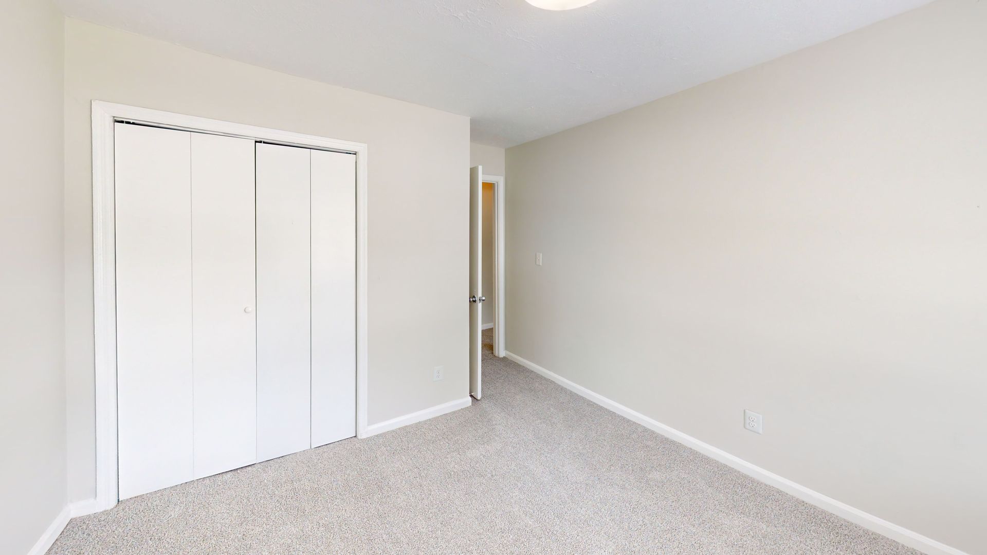 Empty beige bedroom with white closet doors and gray carpet, viewed from the corner.