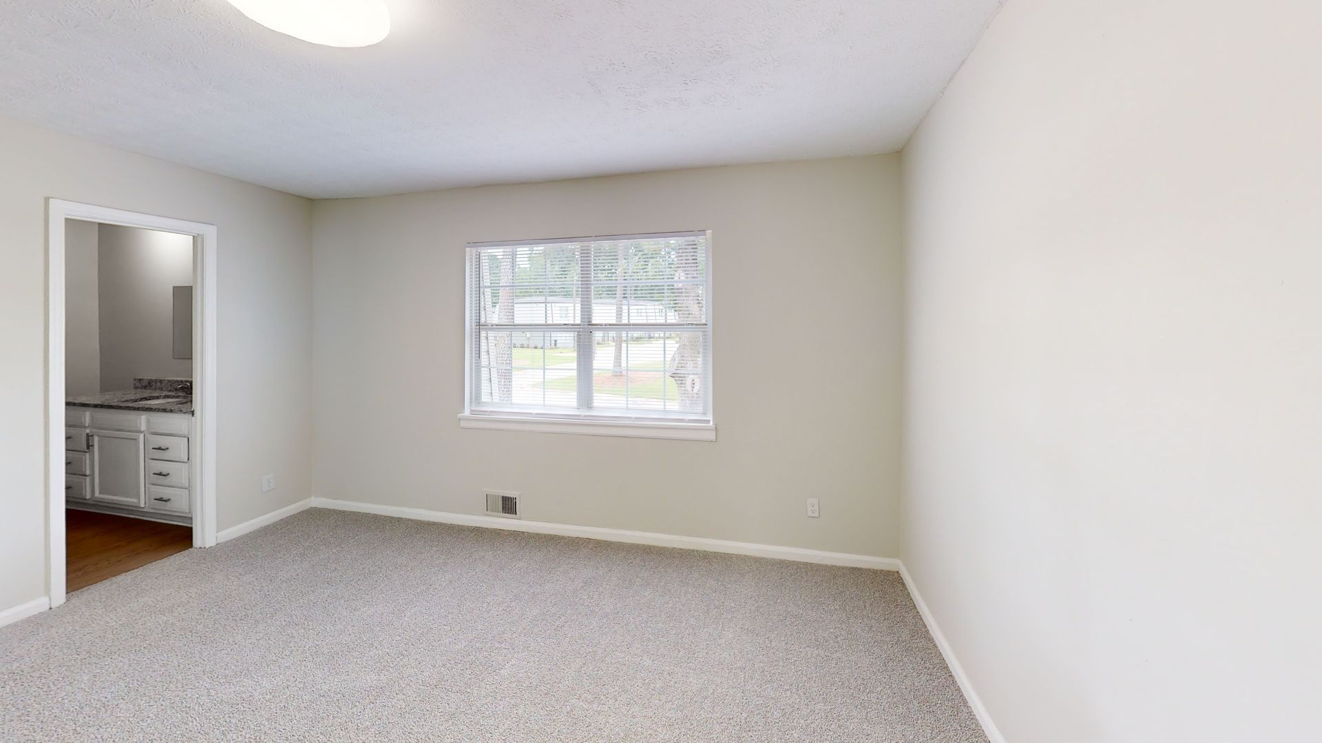 Empty beige bedroom with carpet, window, and open doorway to a bathroom.