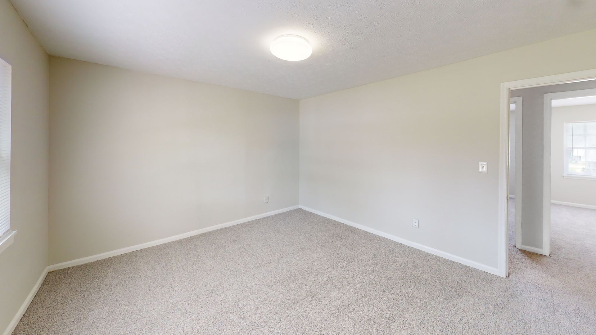 Empty beige bedroom with carpet, window, ceiling light, and open doorway