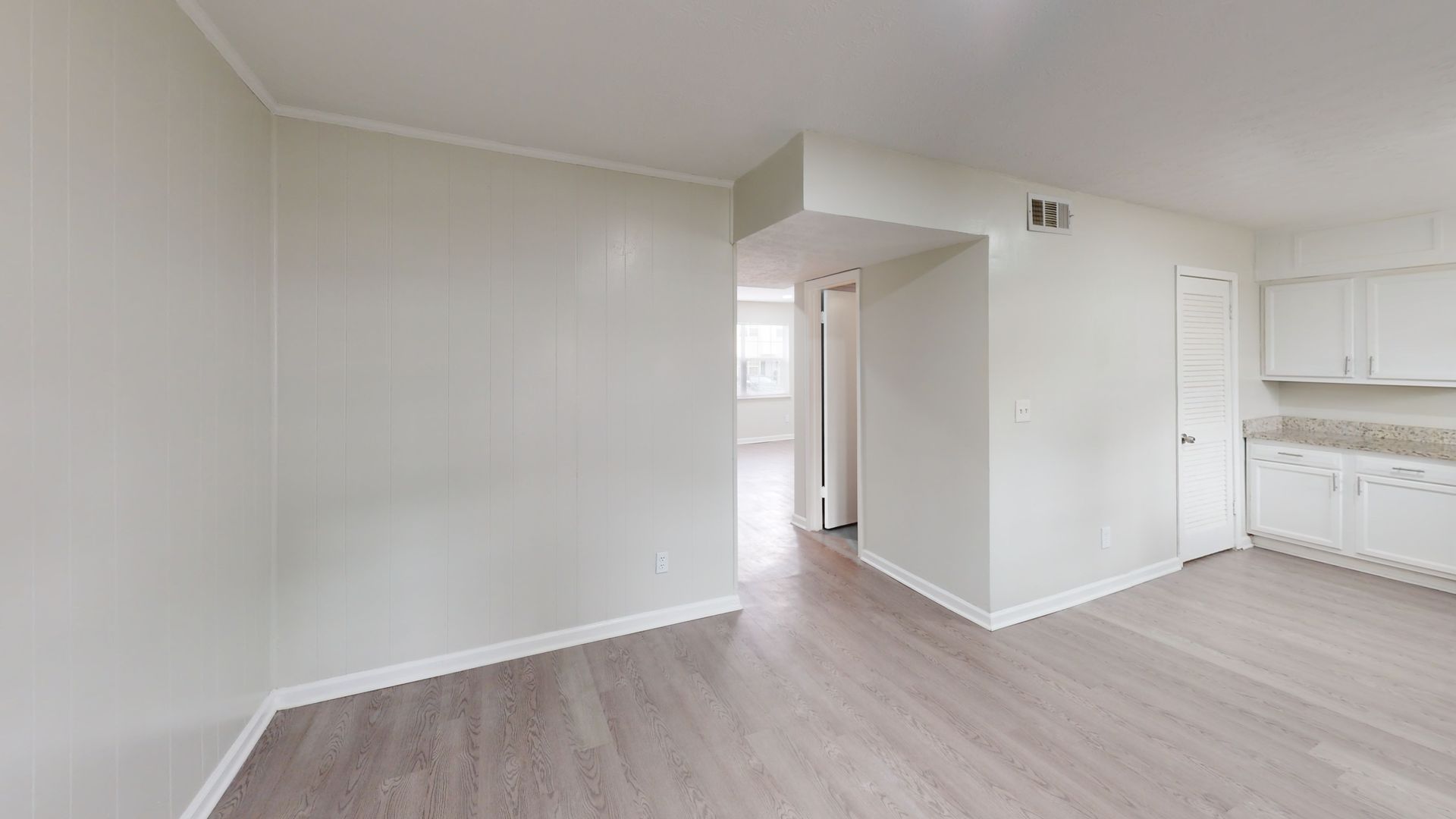 Empty modern room with light gray walls, wood flooring, and a small kitchen area on the right
