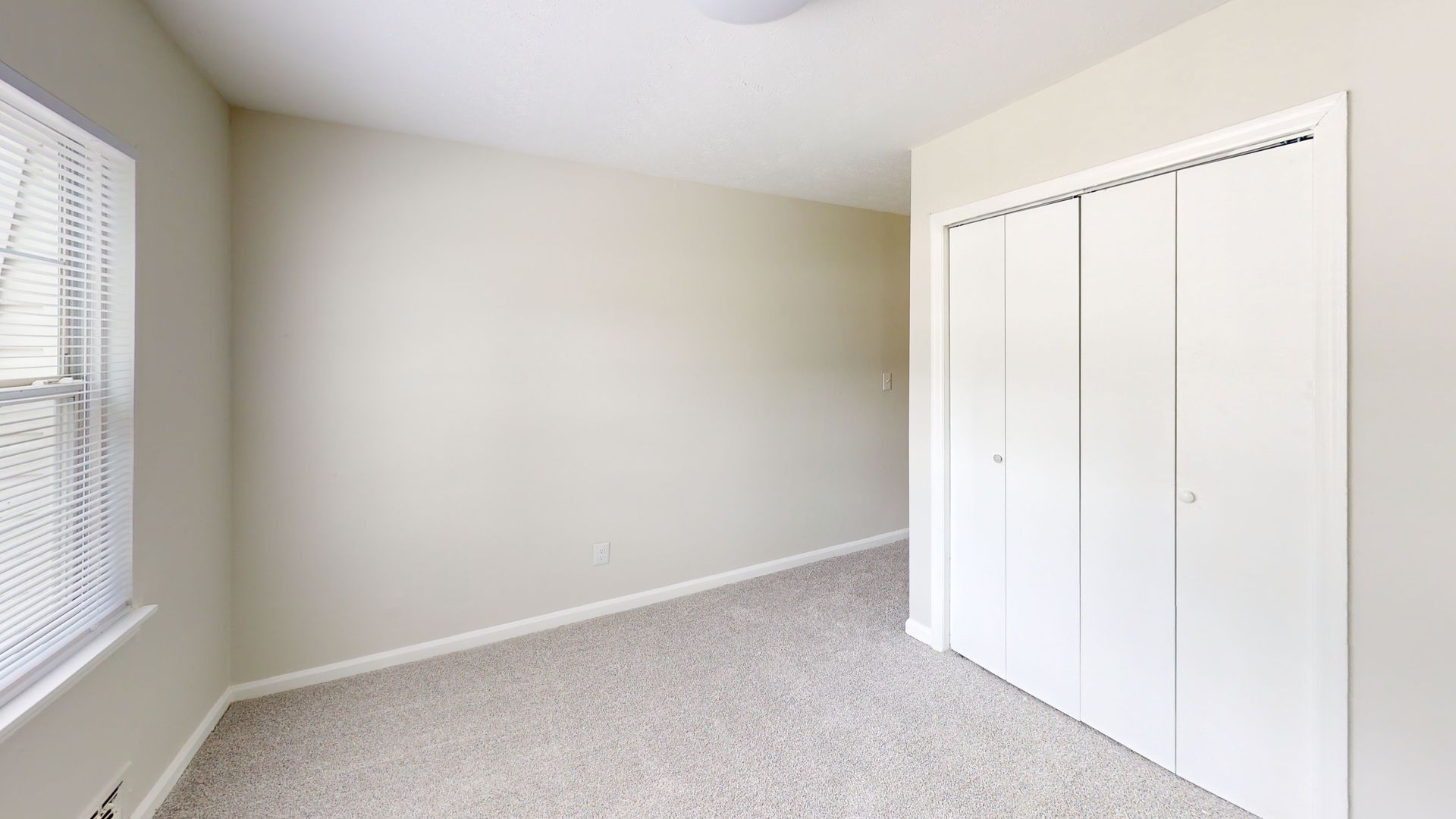 Empty beige bedroom with carpet, window blinds, and white closet doors.