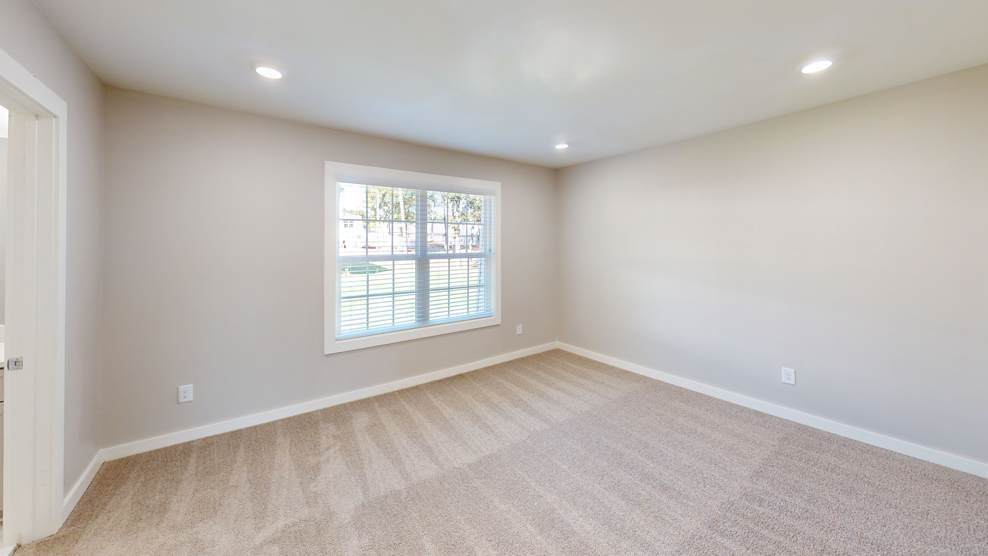 Empty beige bedroom with carpet, recessed lights, and a window letting in natural light