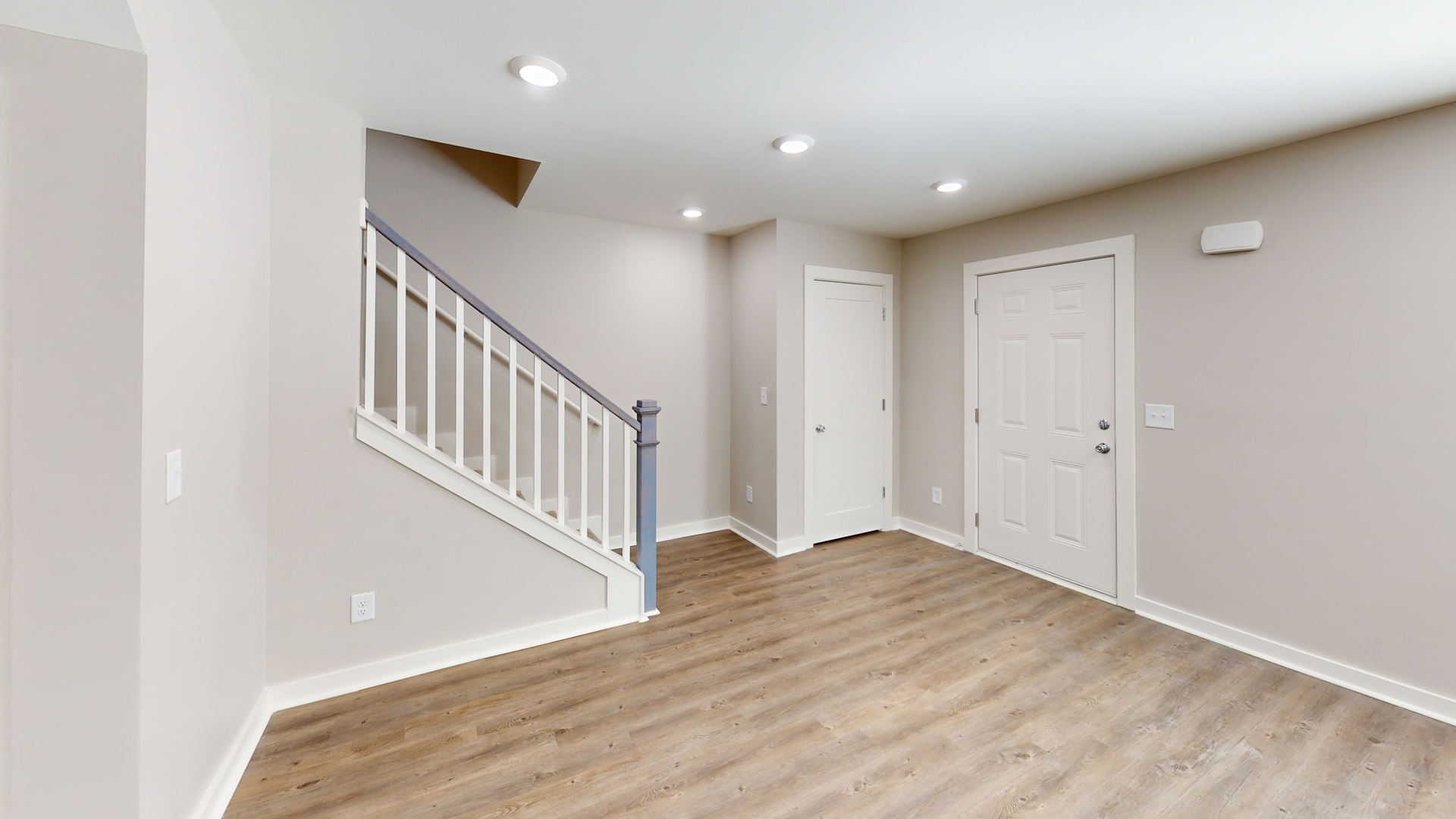 Empty beige hallway with wood floors, white staircase railing, and closed doors under recessed lights