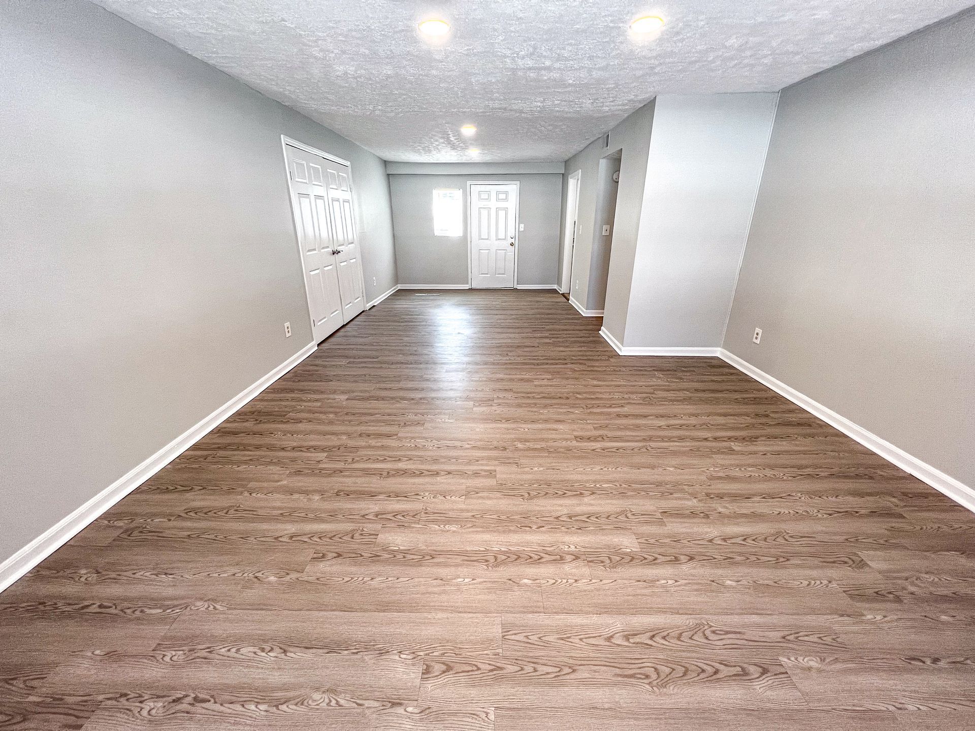 Empty living room with light walls, wood-look floor, and bright windows at the far end