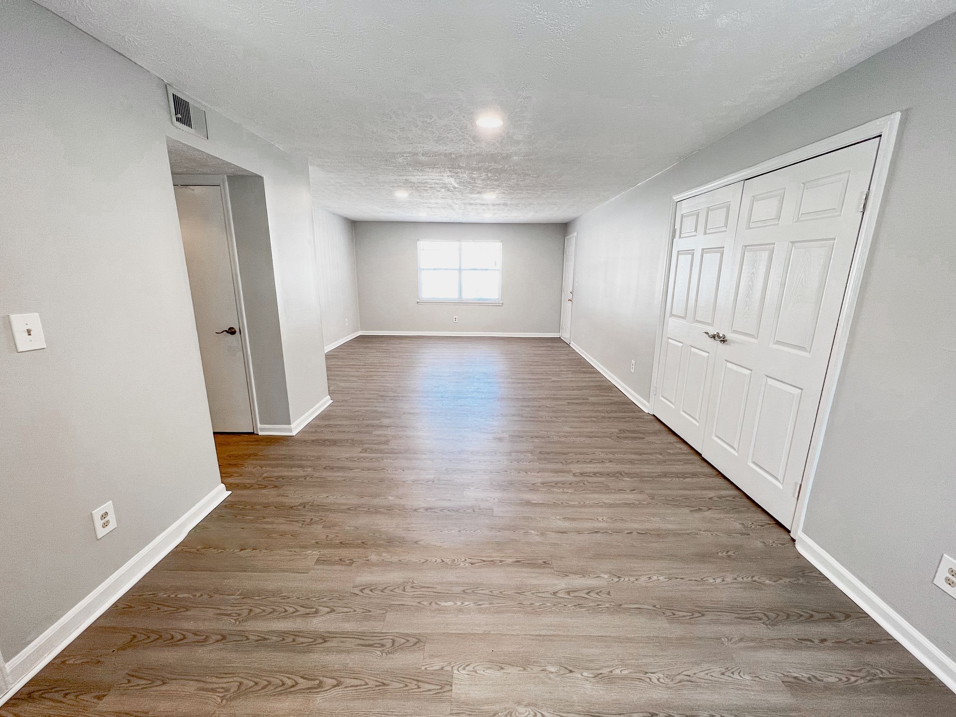 Empty living room with light gray walls, wood flooring, and a window at the far end.