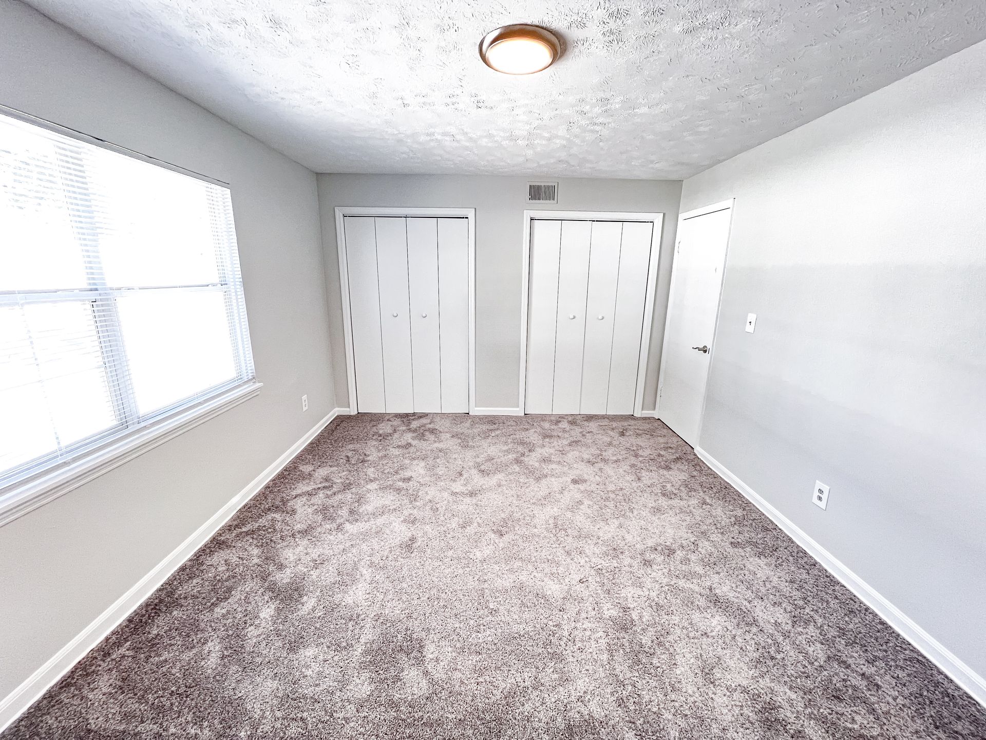 Empty white bedroom with carpet, two closet doors, and a window on the left.