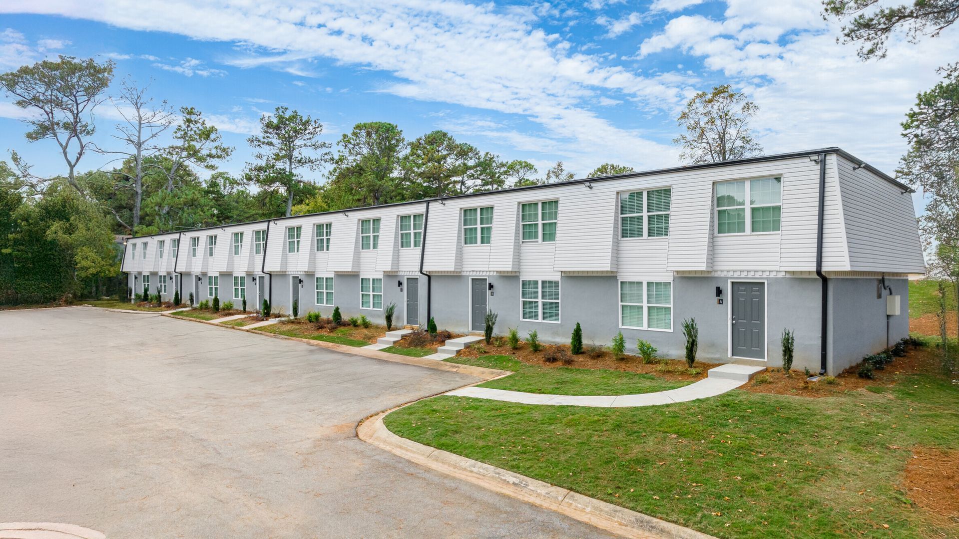 Row of white two-story townhouses beside a driveway and grassy area under a cloudy sky