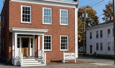 Brick building with white trim and porch, beside a white house on a quiet street.