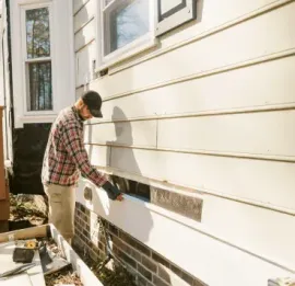 Person painting the exterior of a house with white siding and brick trim.
