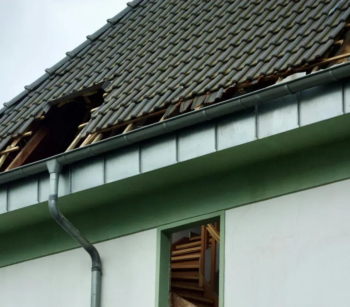 Damaged house roof with missing tiles and open attic window above a green wall