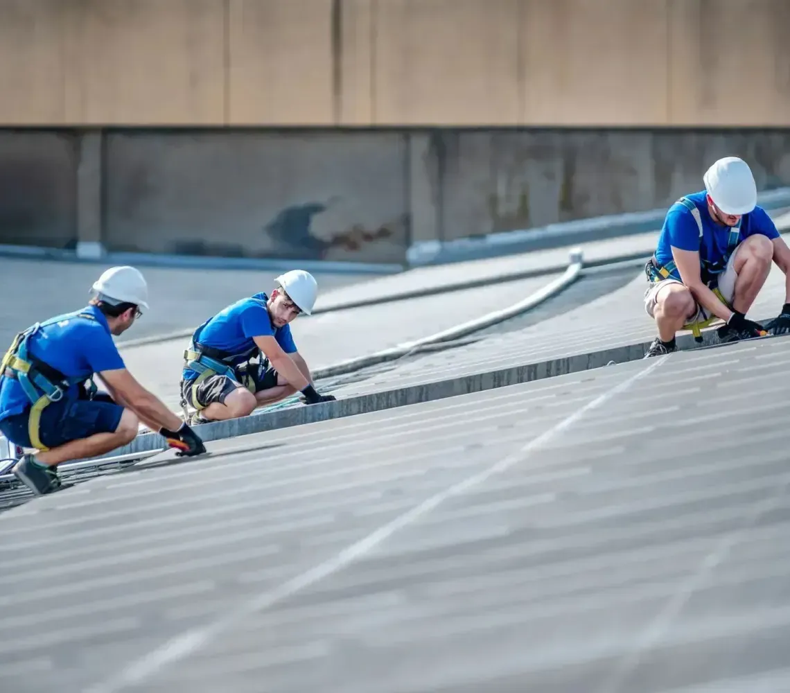 Three workers in blue safety gear install roofing on a steep metal roof