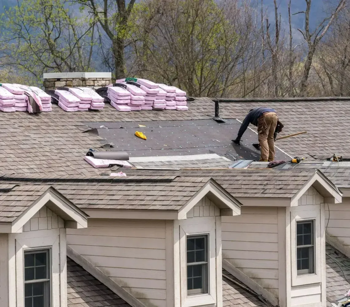 Two workers installing shingles on a house roof with stacks of roofing materials nearby.