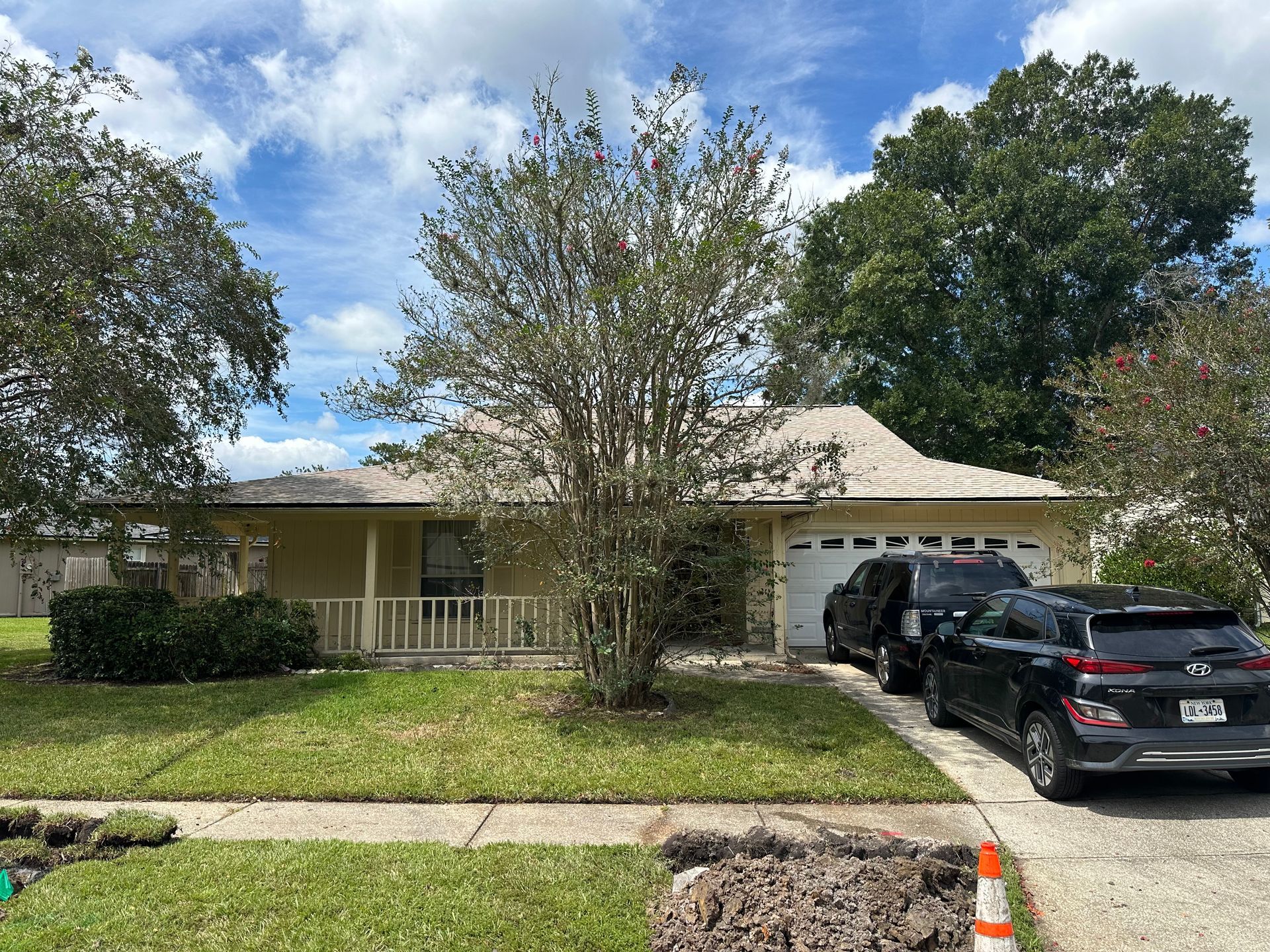 Single-story suburban house with a front lawn, tree, and two cars parked in the driveway.