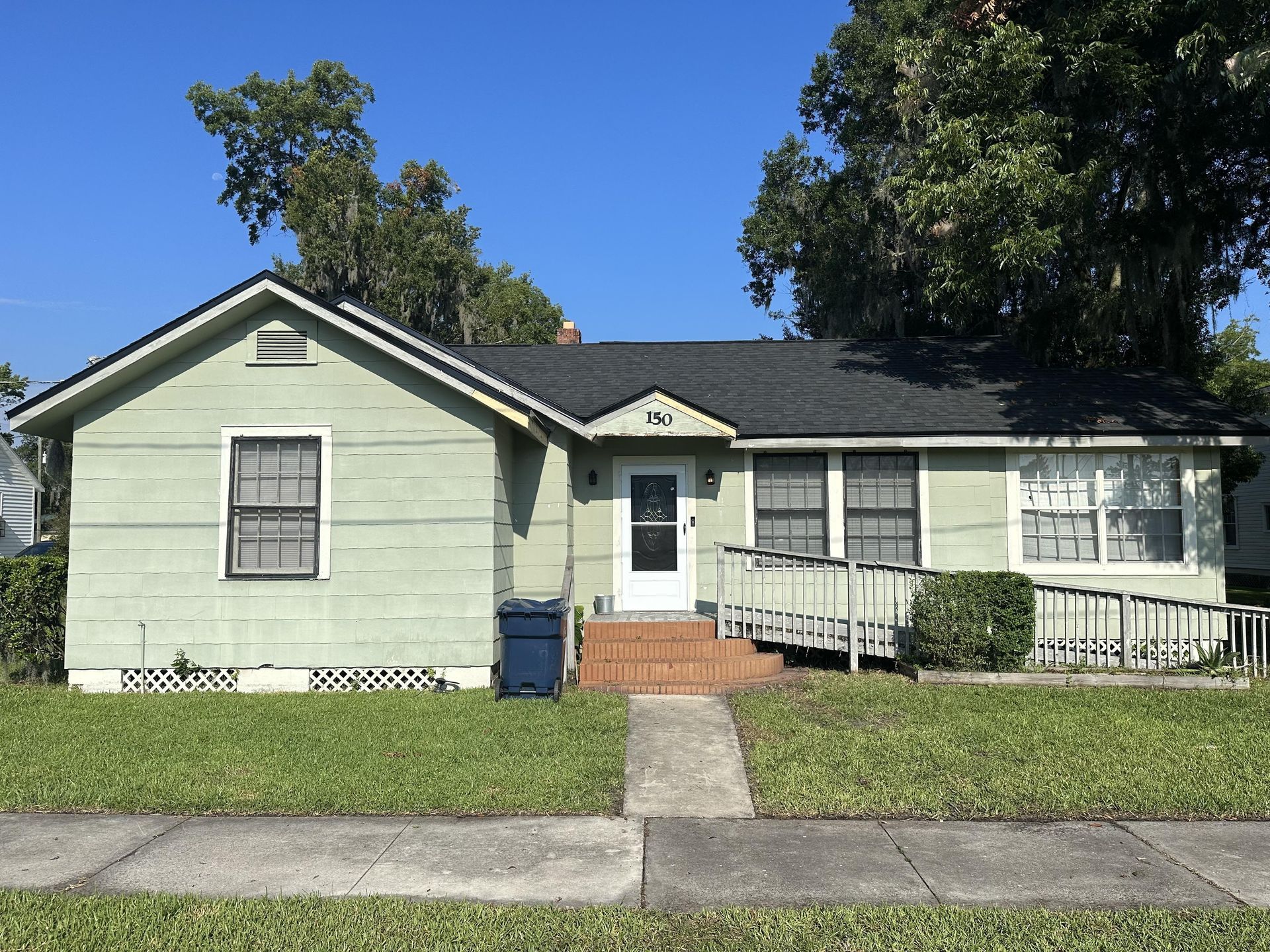 Small pale green bungalow with front steps, black roof, and a wheelchair ramp under a blue sky.