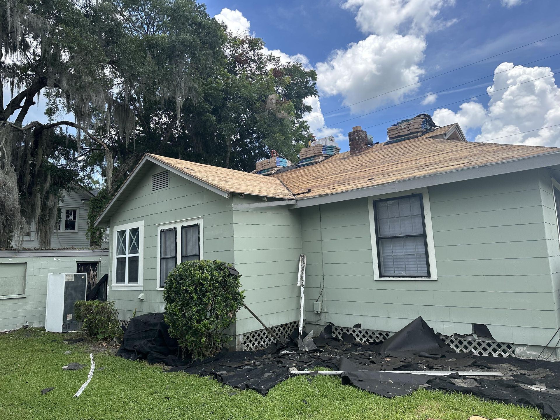 Single-story light green house with damaged roof shingles and debris in the yard after storm damage