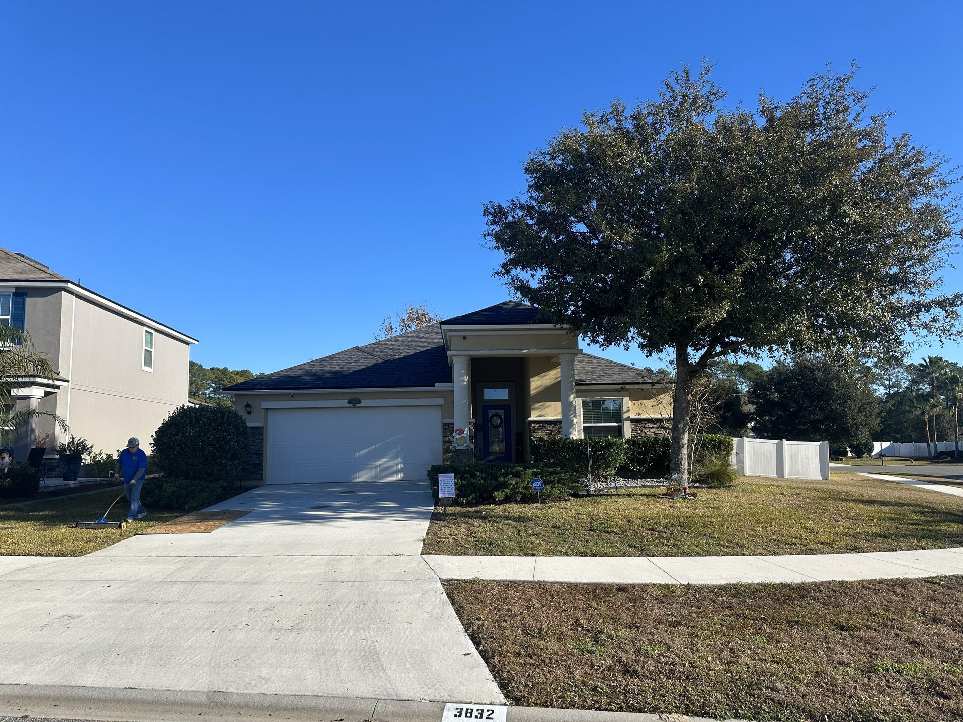 Suburban single-story house with two-car garage, driveway, and large front tree under a clear blue sky