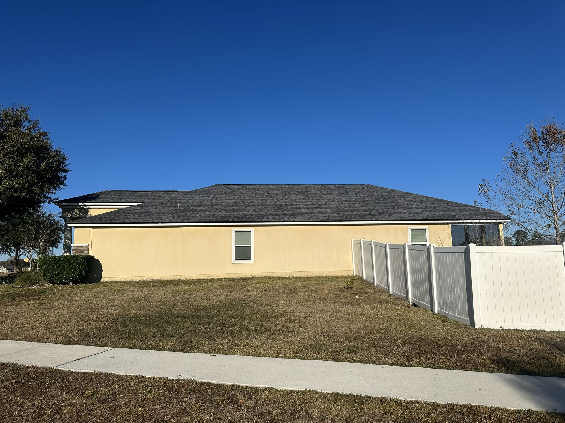 Single-story beige house with dark roof, fenced side yard, and a sidewalk under a clear blue sky