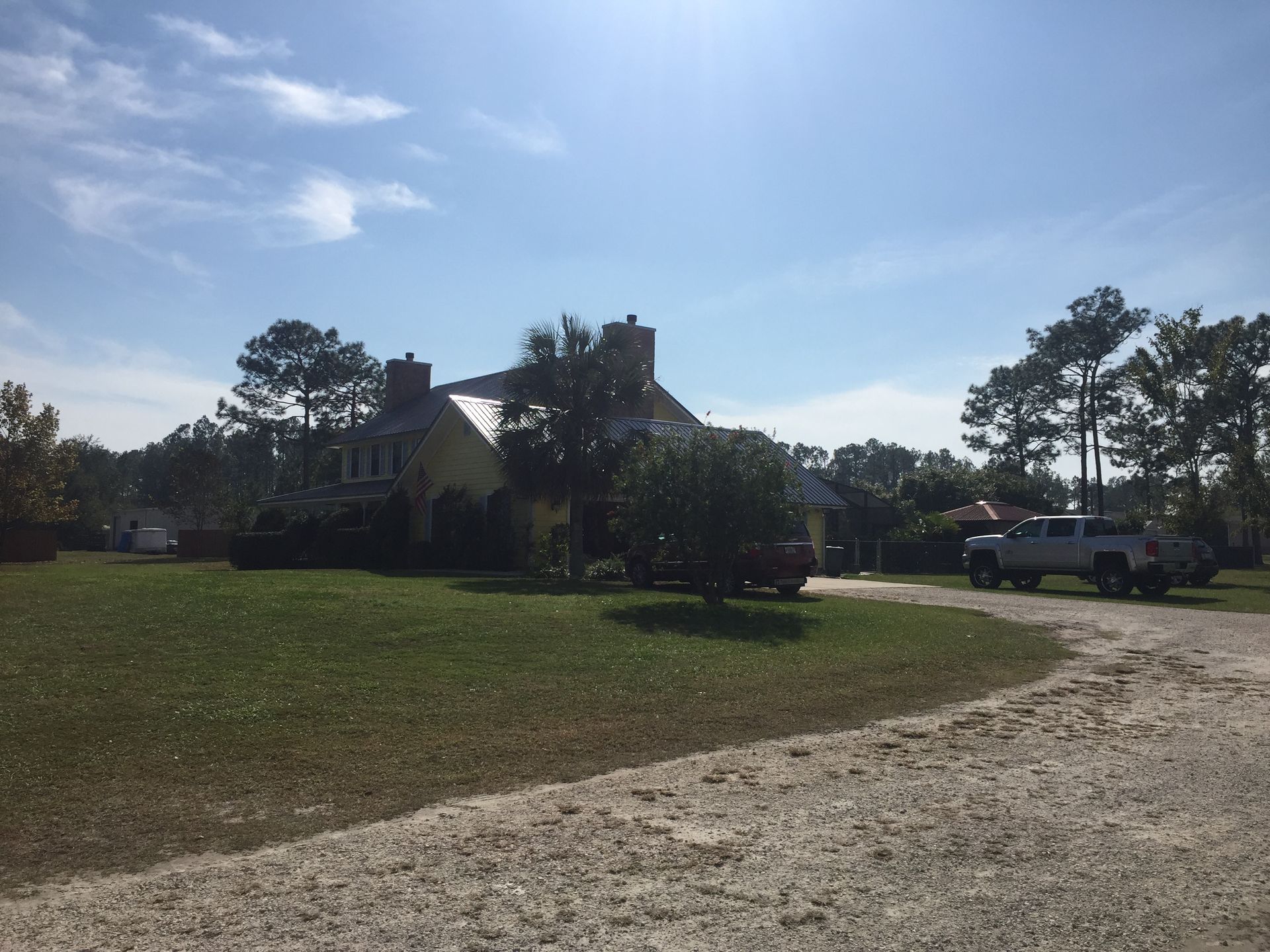 Rural house with a pickup truck on a gravel driveway, surrounded by trees under a bright blue sky.