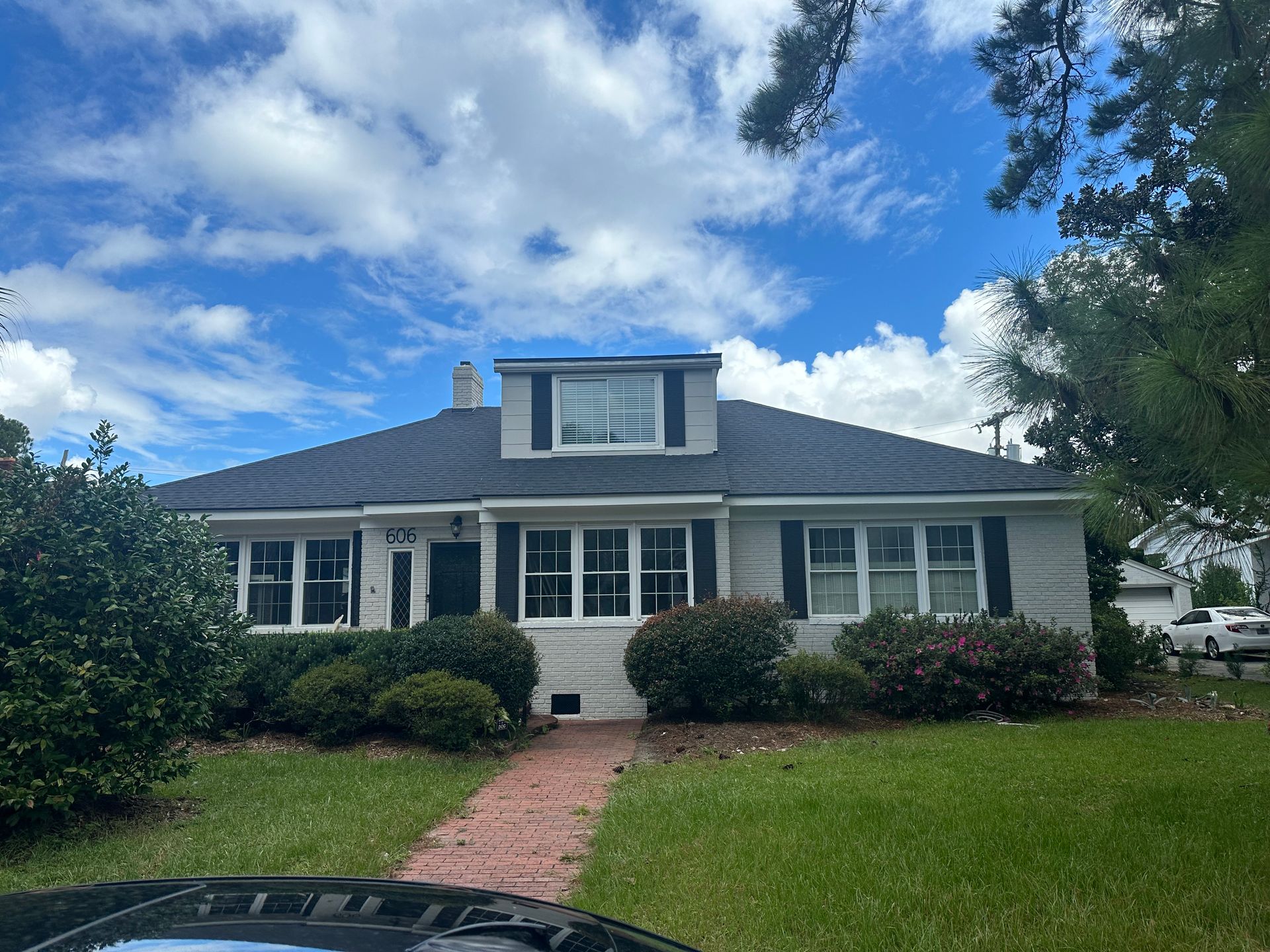 Single-story white house with dormer, dark roof, red brick walkway, and green lawn under a blue sky