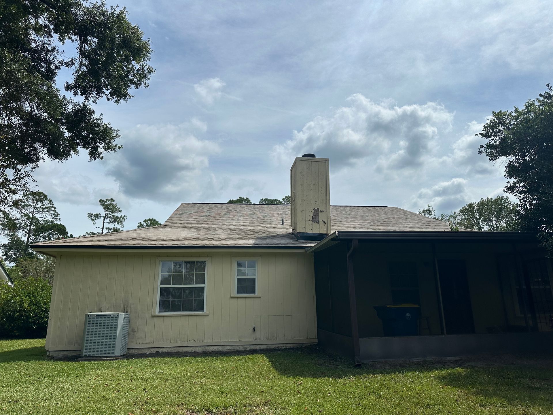 Small beige house with a chimney and covered porch under a cloudy sky, framed by trees.