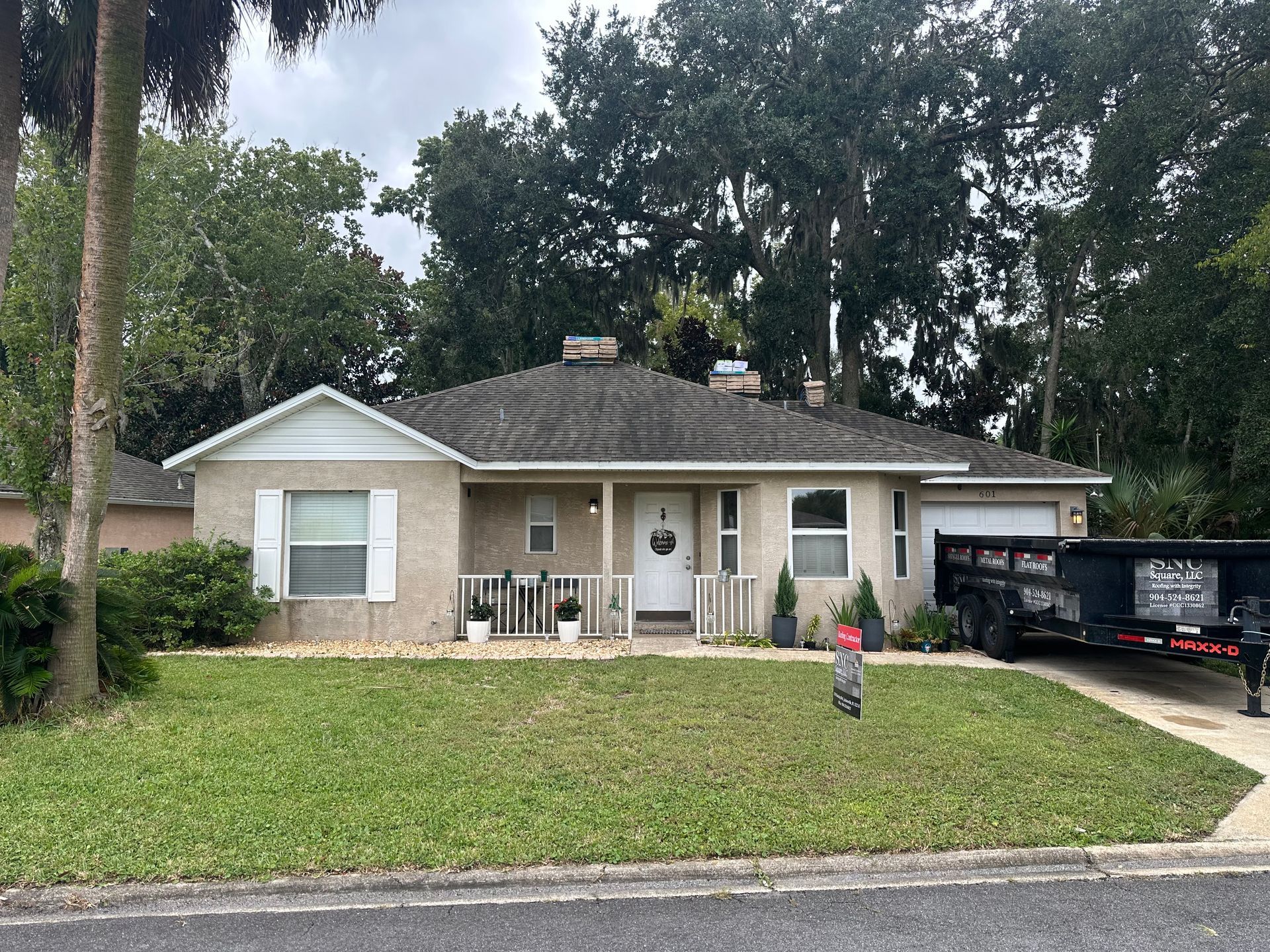 Single-story beige house with white shutters, front porch, and a trailer in the driveway under tall trees