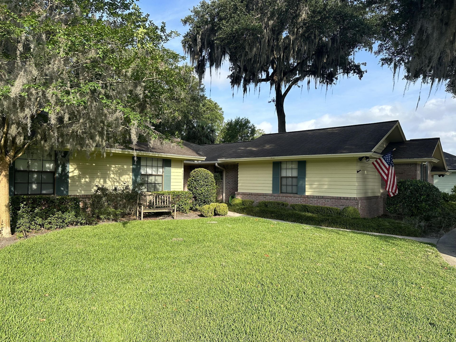 Single-story cream house with green shutters, a large front lawn, and an American flag by the porch.