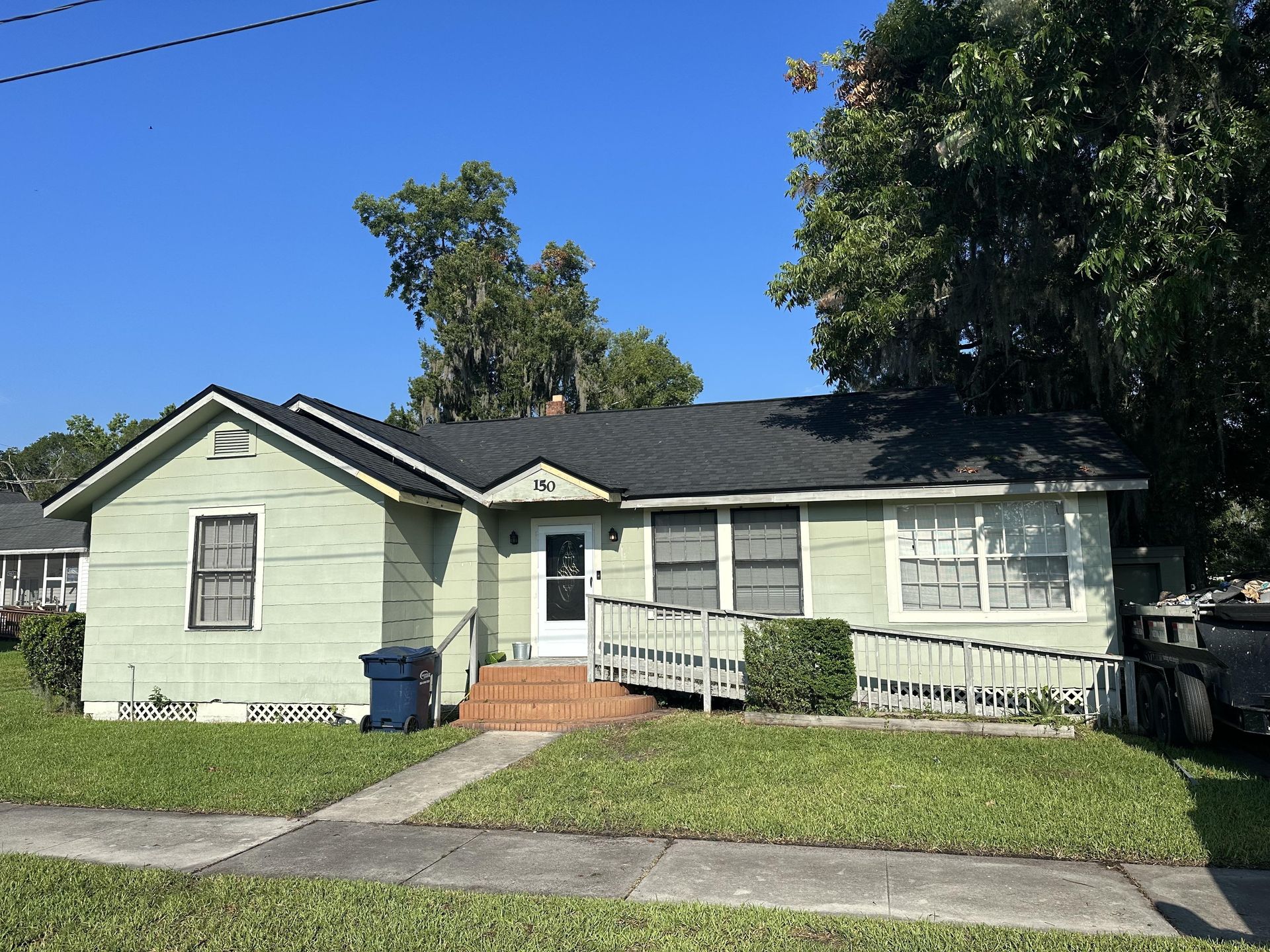 Small light-green house with a ramp, front steps, and a blue bin under a clear blue sky
