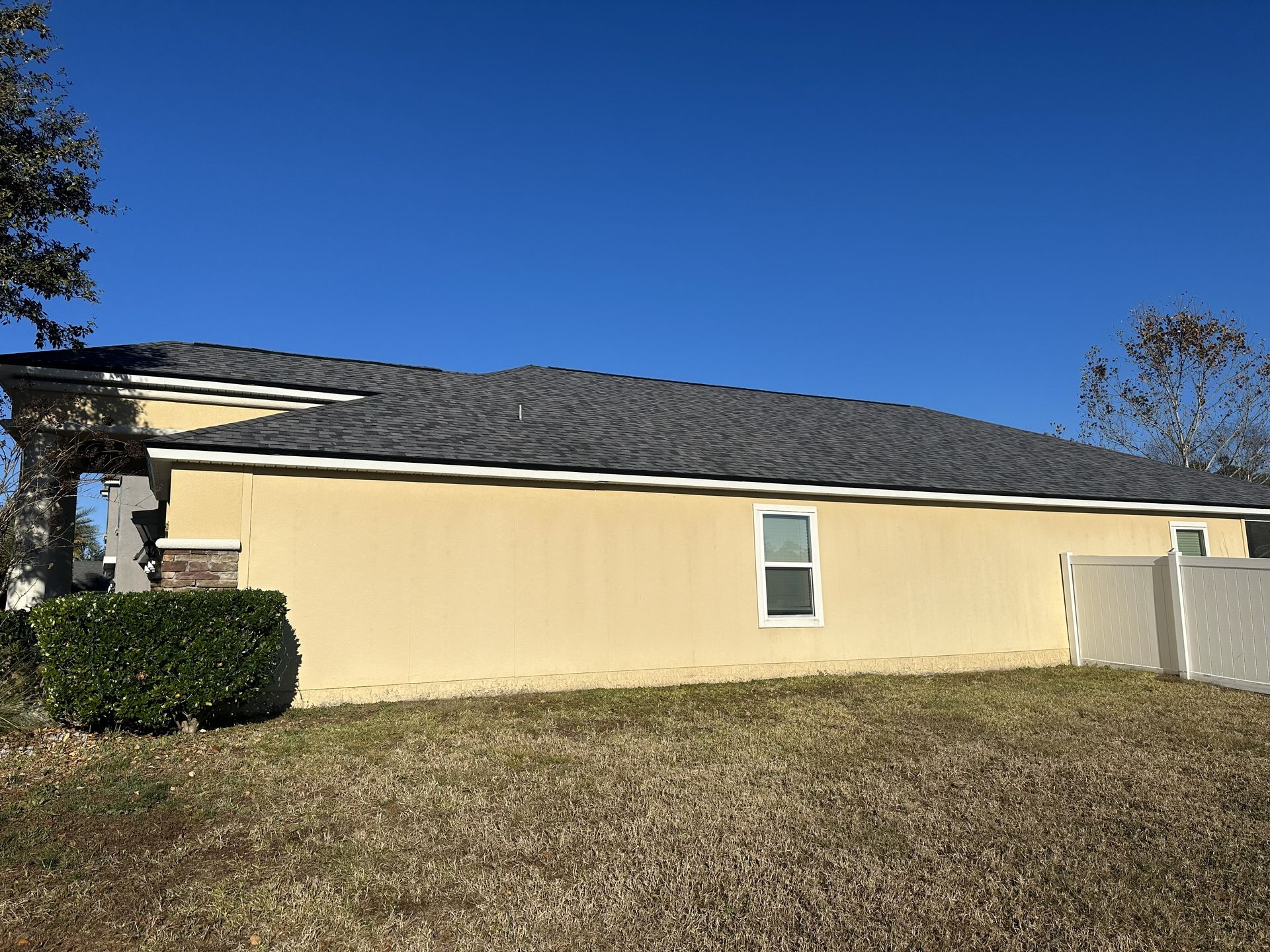 Single-story beige house with dark roof and small window, seen across a dry grassy yard under a blue sky
