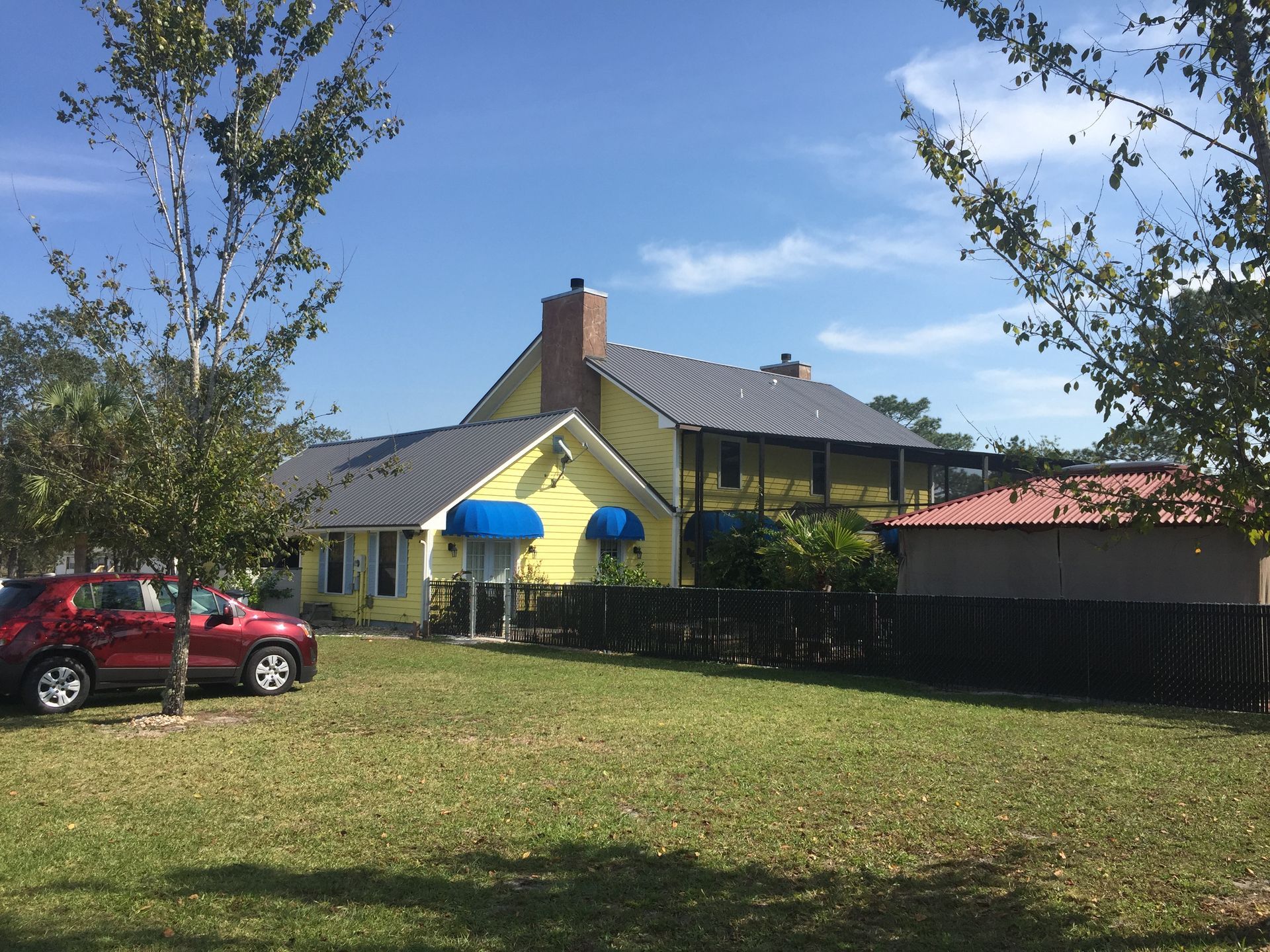 Yellow house with blue awnings, red car, and fenced yard on a sunny day