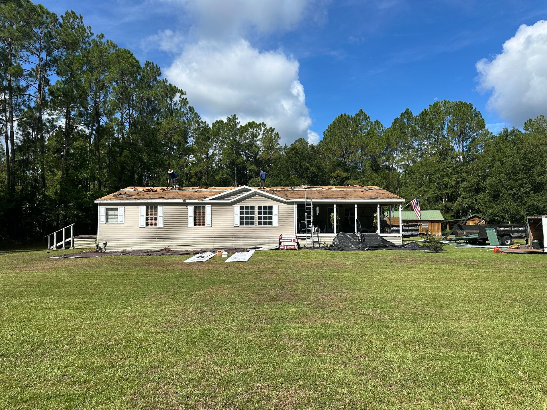 Single-story beige house with a damaged roof, front porch, and large grassy yard under a blue sky