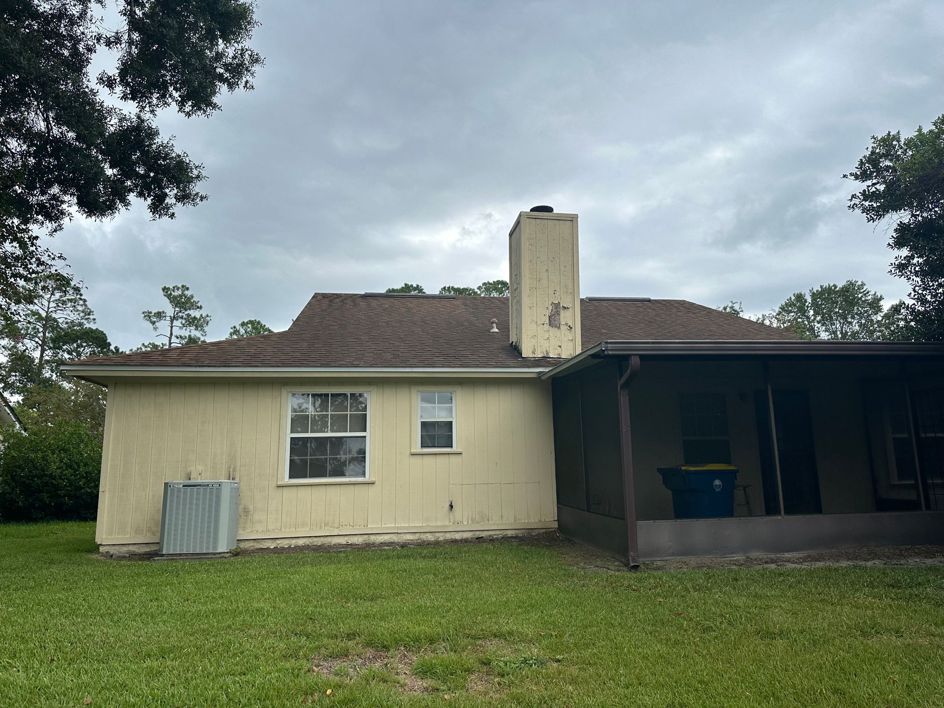 Backyard view of a beige house with brown roof, screened porch, and grassy lawn under cloudy sky