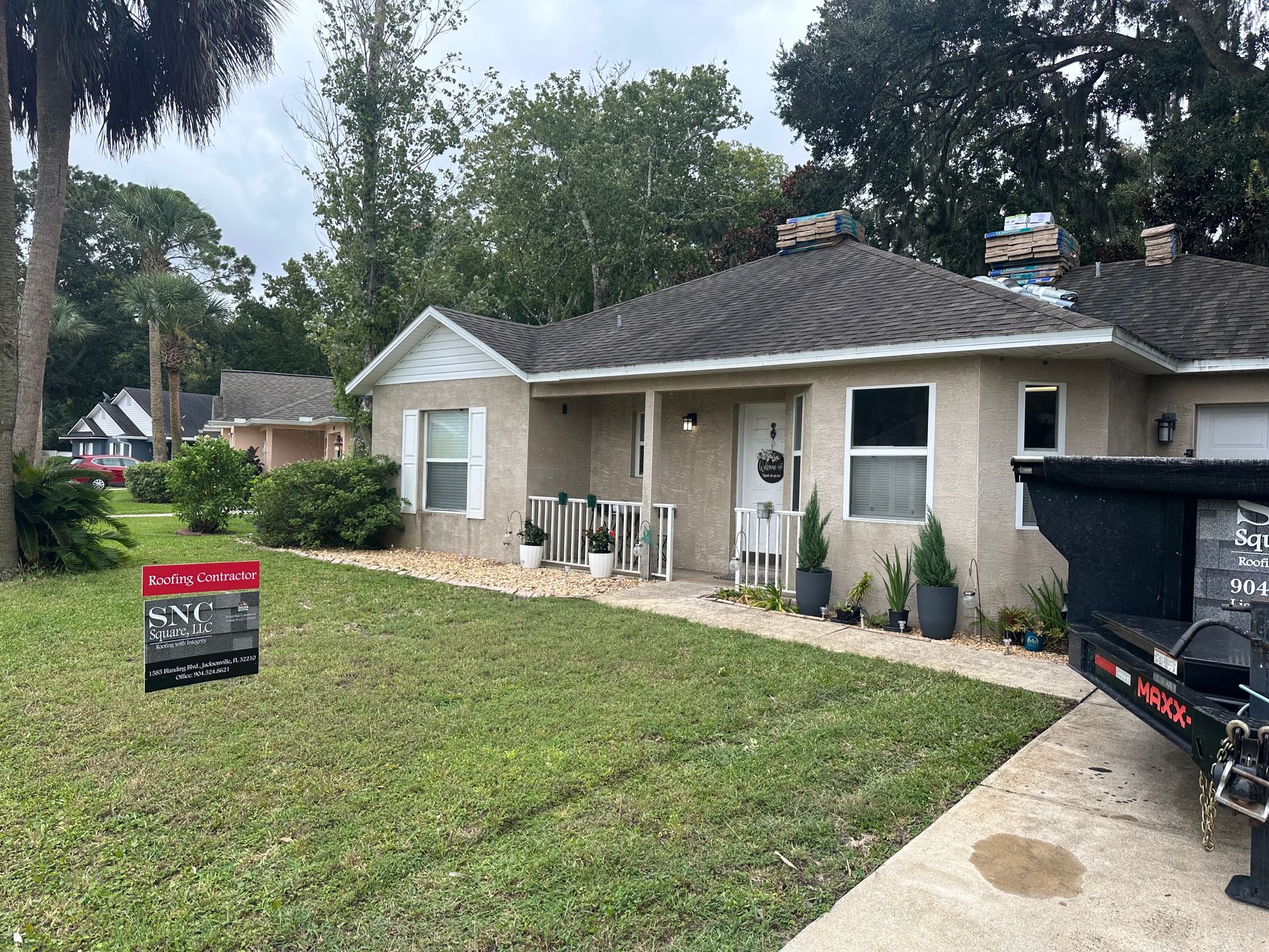 Single-story beige house with a front lawn, driveway, patio chairs, and a black dumpster in the foreground