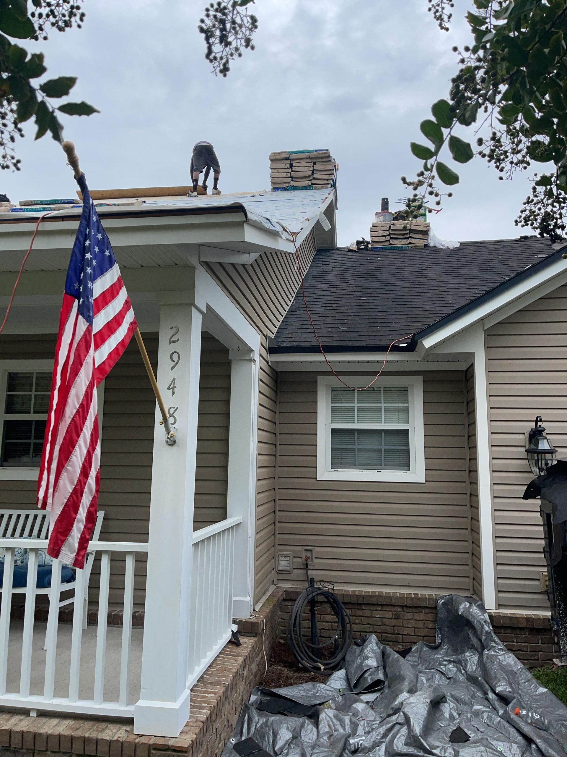 House with severe roof damage, tarped debris below, and an American flag on the porch.