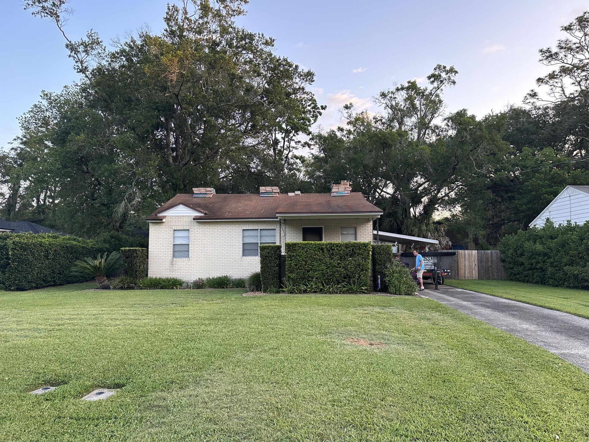 Small white house with a red roof beside a grassy yard and driveway, surrounded by trees.