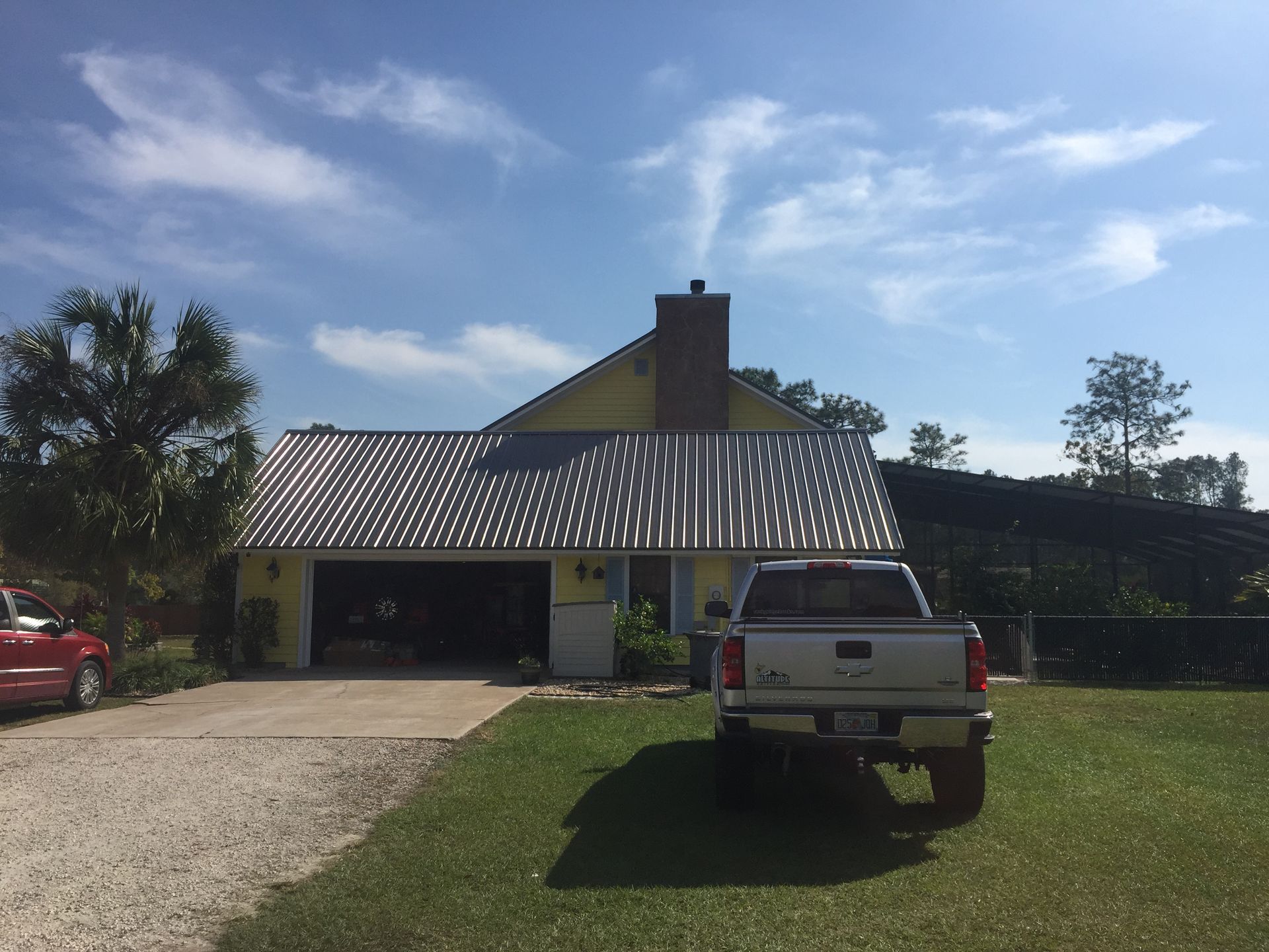 Rural house with metal roof, garage, and white pickup truck parked on a grassy yard under a blue sky.