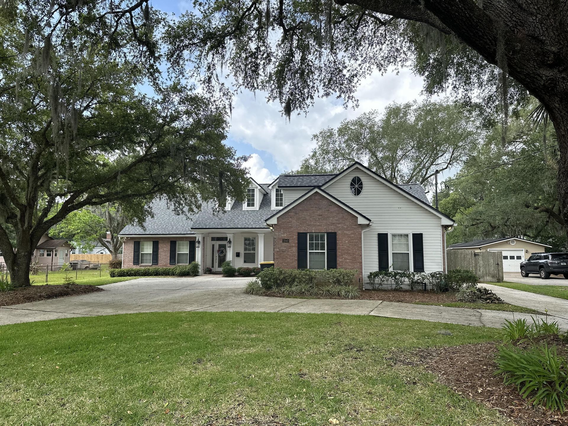 Suburban brick-and-white house with a front lawn, driveway, and large trees overhead