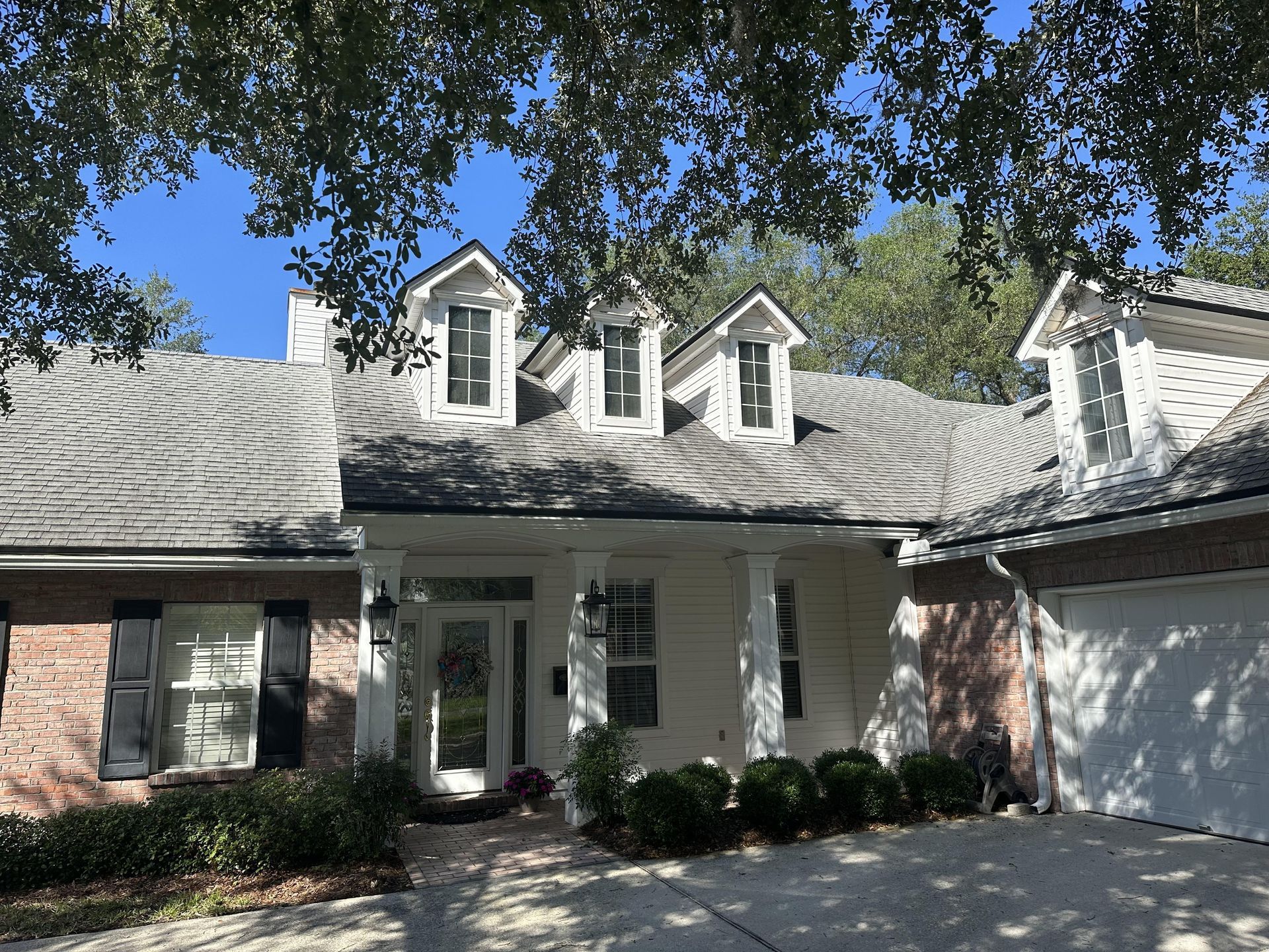 Brick house exterior with gray roof, dormer windows, white garage, and front entry under trees
