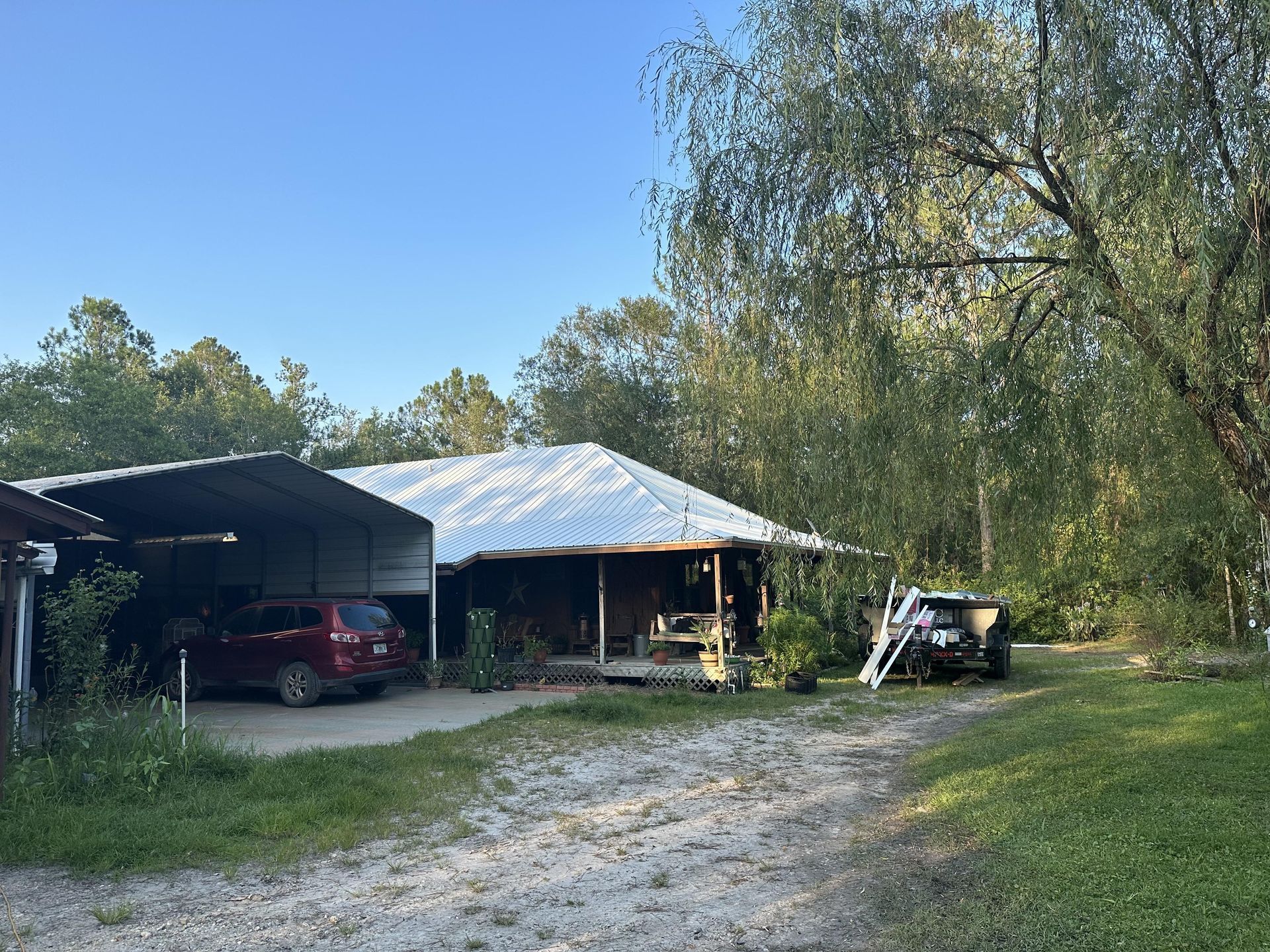 Rural yard with a carport, red car, gravel driveway, and trees under a clear blue sky