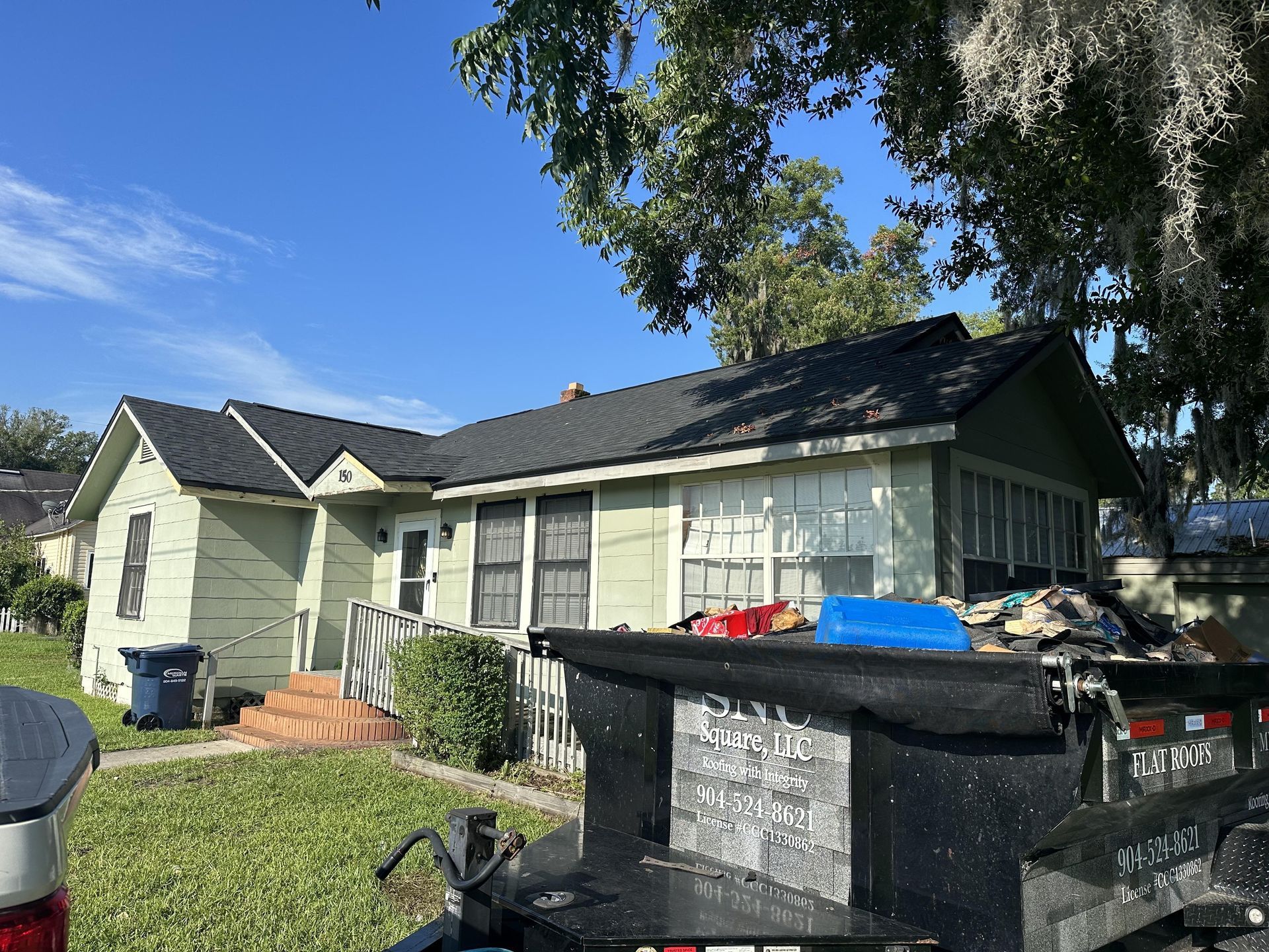 Single-story house with a green lawn, trash bins, and a large black dumpster in the front yard