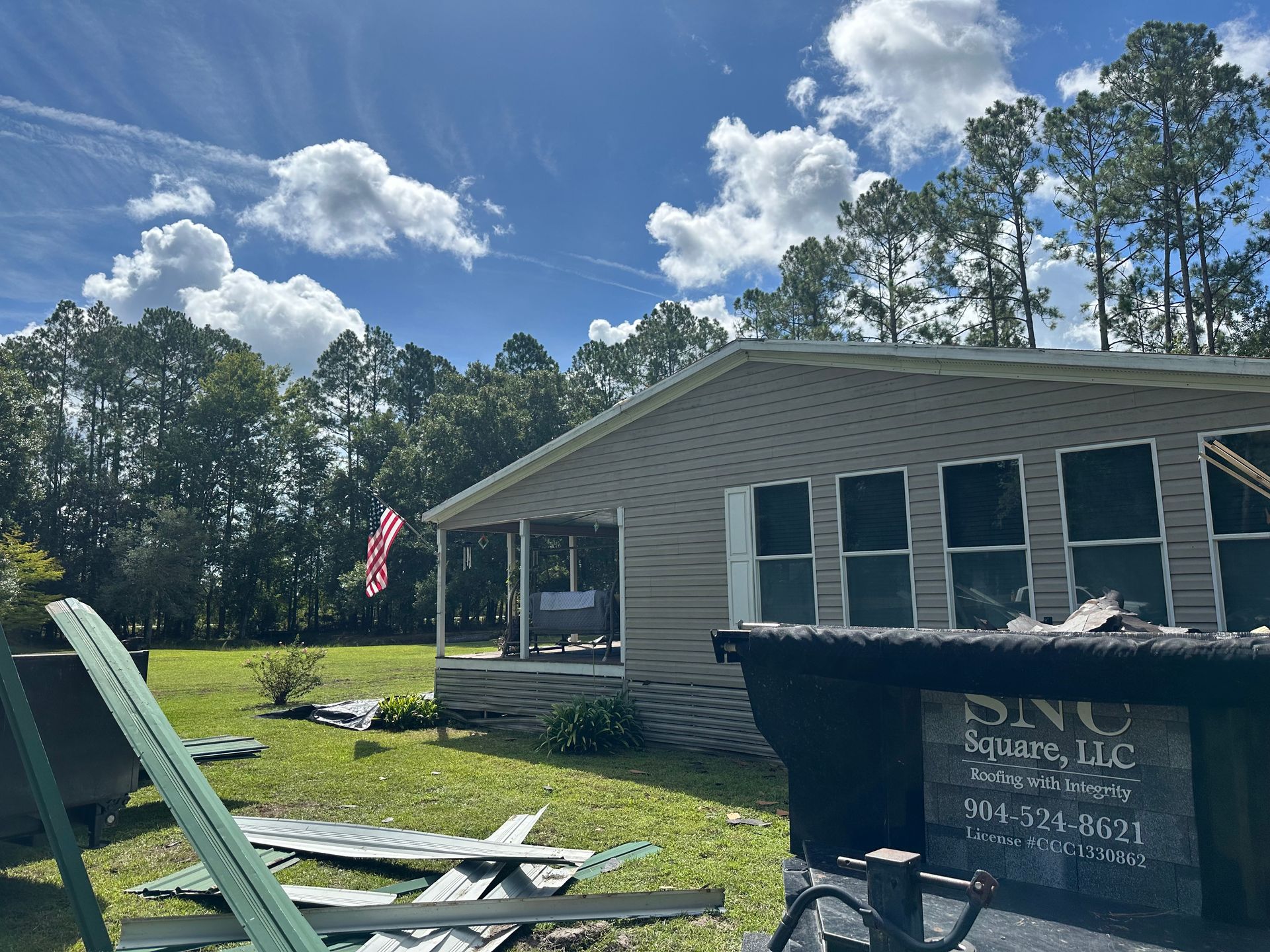 Single-story house with green shutters and porch, American flag, and construction debris in the yard under a blue sky