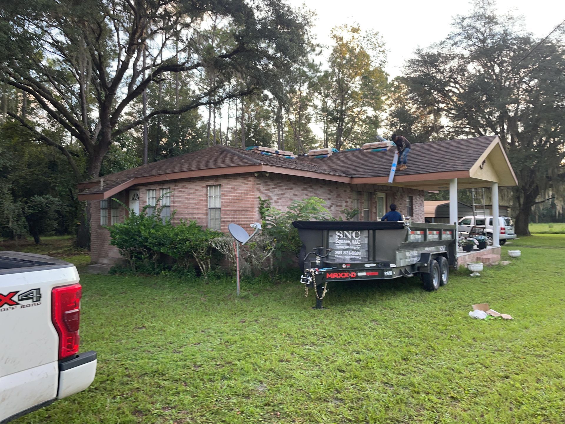 Brick house with damaged roof, utility trailer, and pickup truck on a grassy yard after a storm