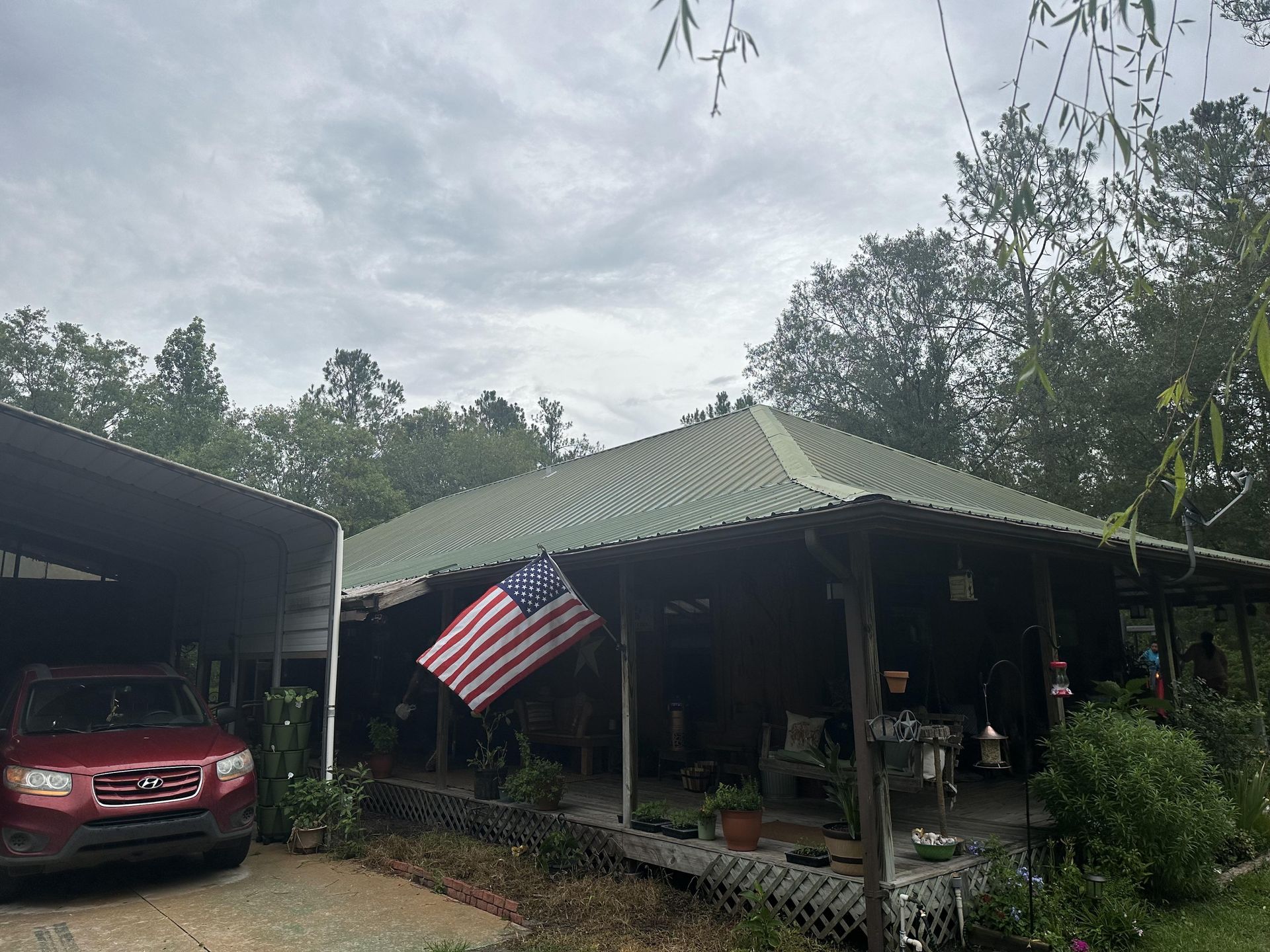 Rural house with a green roof, American flag on the porch, and a red SUV parked under a carport.
