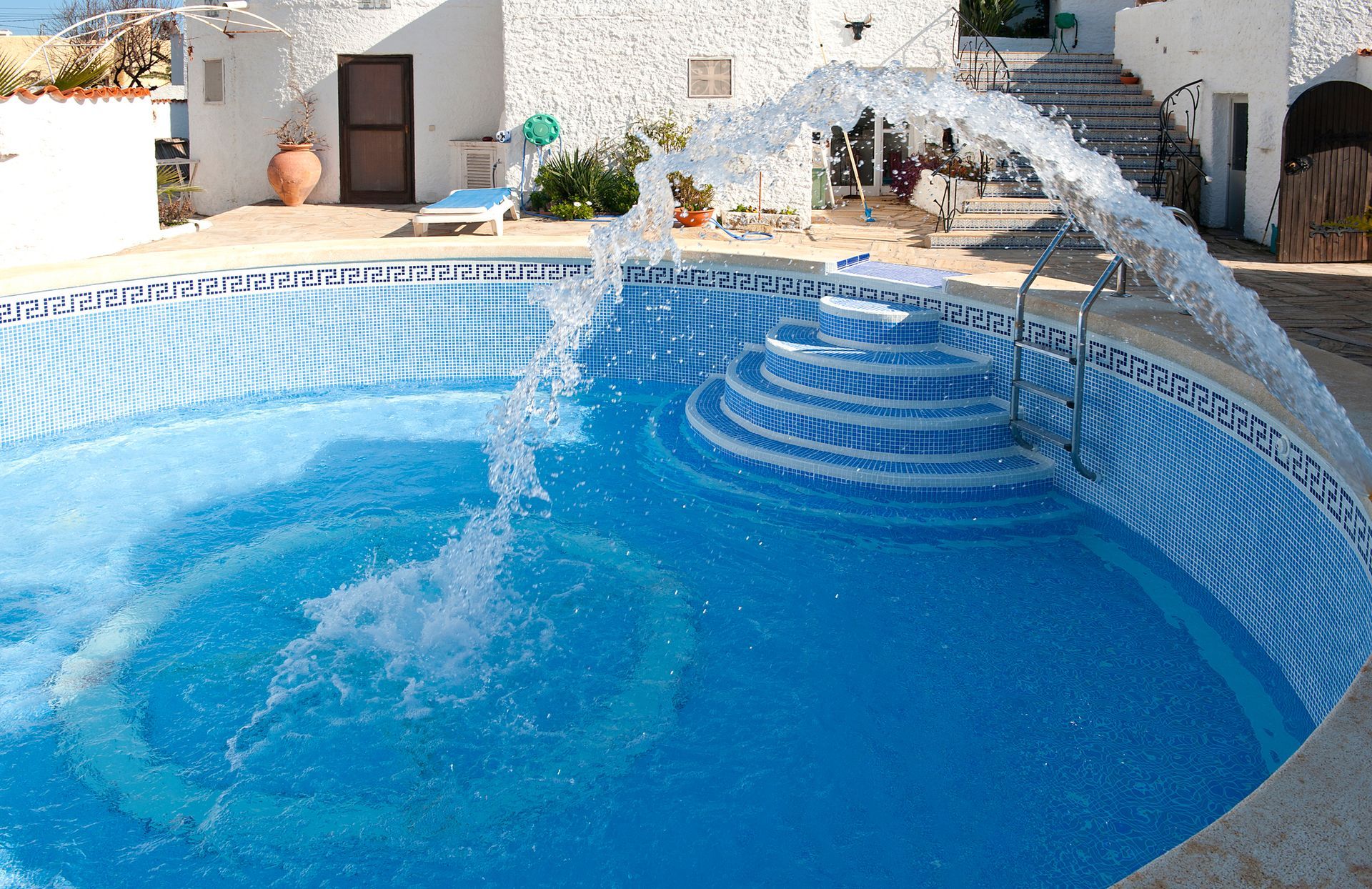 Blue swimming pool with water jets splashing in a sunny white-tiled courtyard