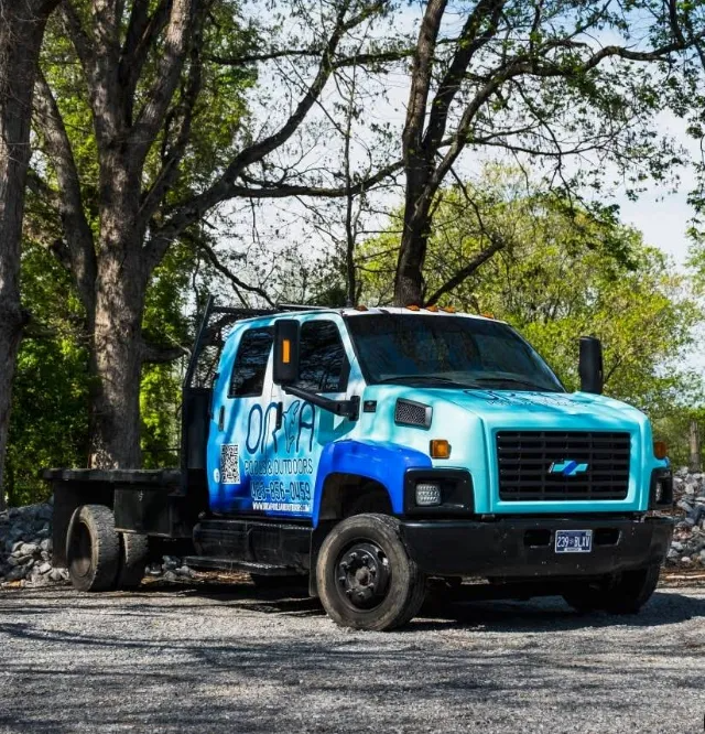 Blue and black tow truck parked under trees on a gravel lot