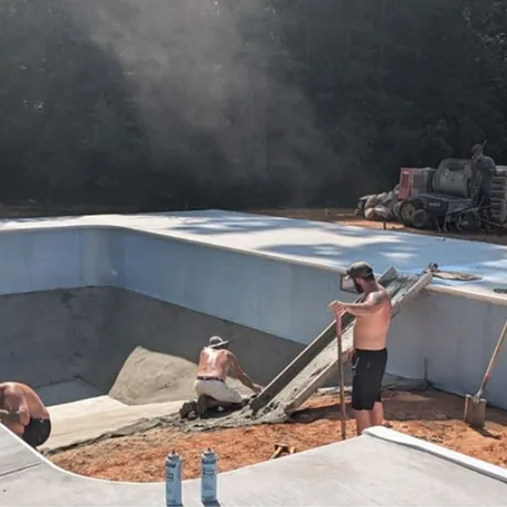 Workers finishing a blue-painted swimming pool with concrete and a cement mixer on a dusty construction site