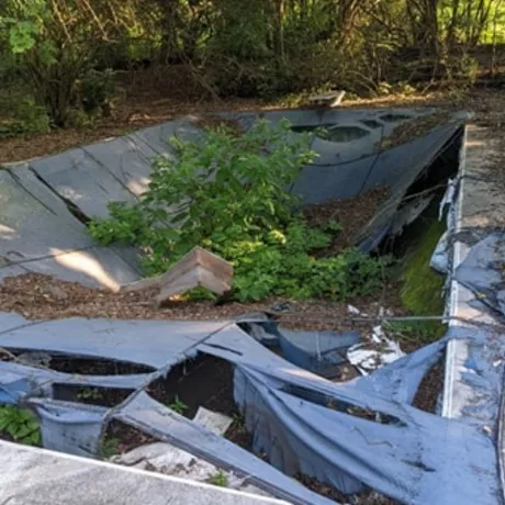 Collapsed blue tarp or liner in a shallow, overgrown pit with debris and greenery.