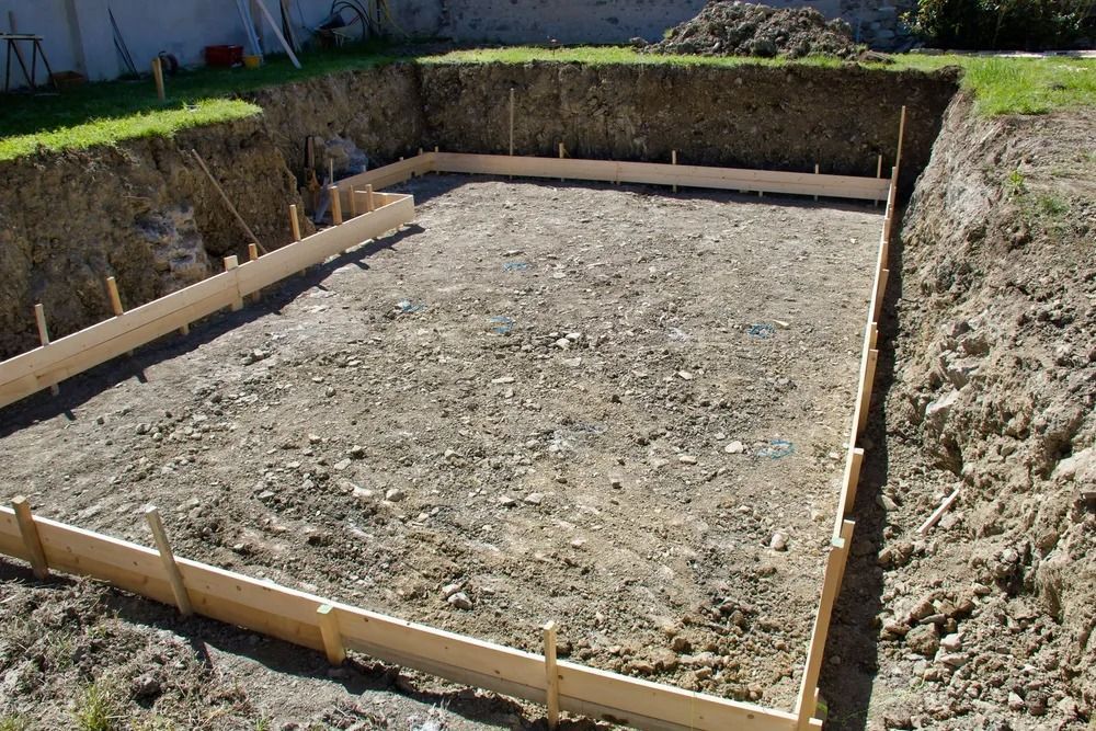 Freshly dug rectangular foundation footing with wooden formwork in a dirt lot